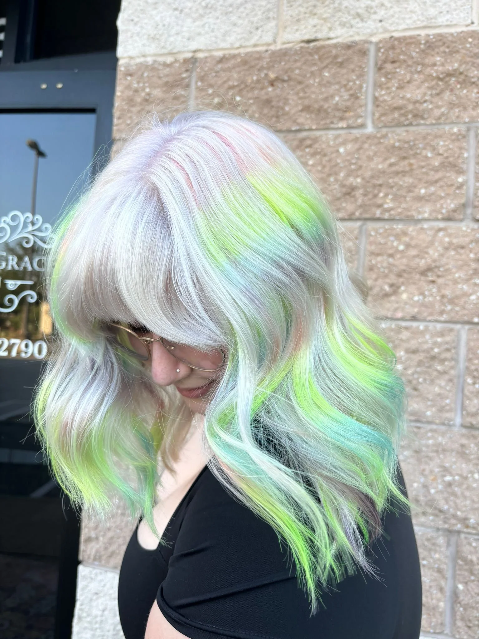 A woman with platinum blonde hair featuring streaks of pink, green, and blue, standing outdoors in front of a brick wall, wearing glasses and a black top.