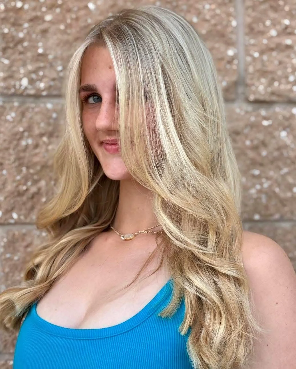 Portrait of a young woman with long, wavy blonde hair, wearing a blue top and a gold necklace, standing against a textured brick wall.