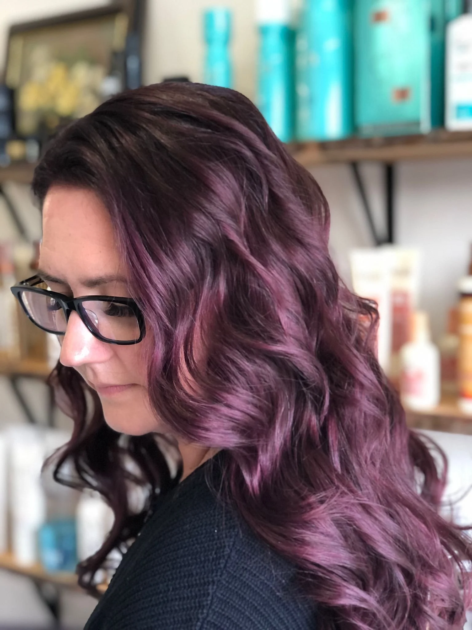 Close-up of a woman with dark wavy hair featuring purple and pink highlights, wearing glasses, in a salon or store setting.