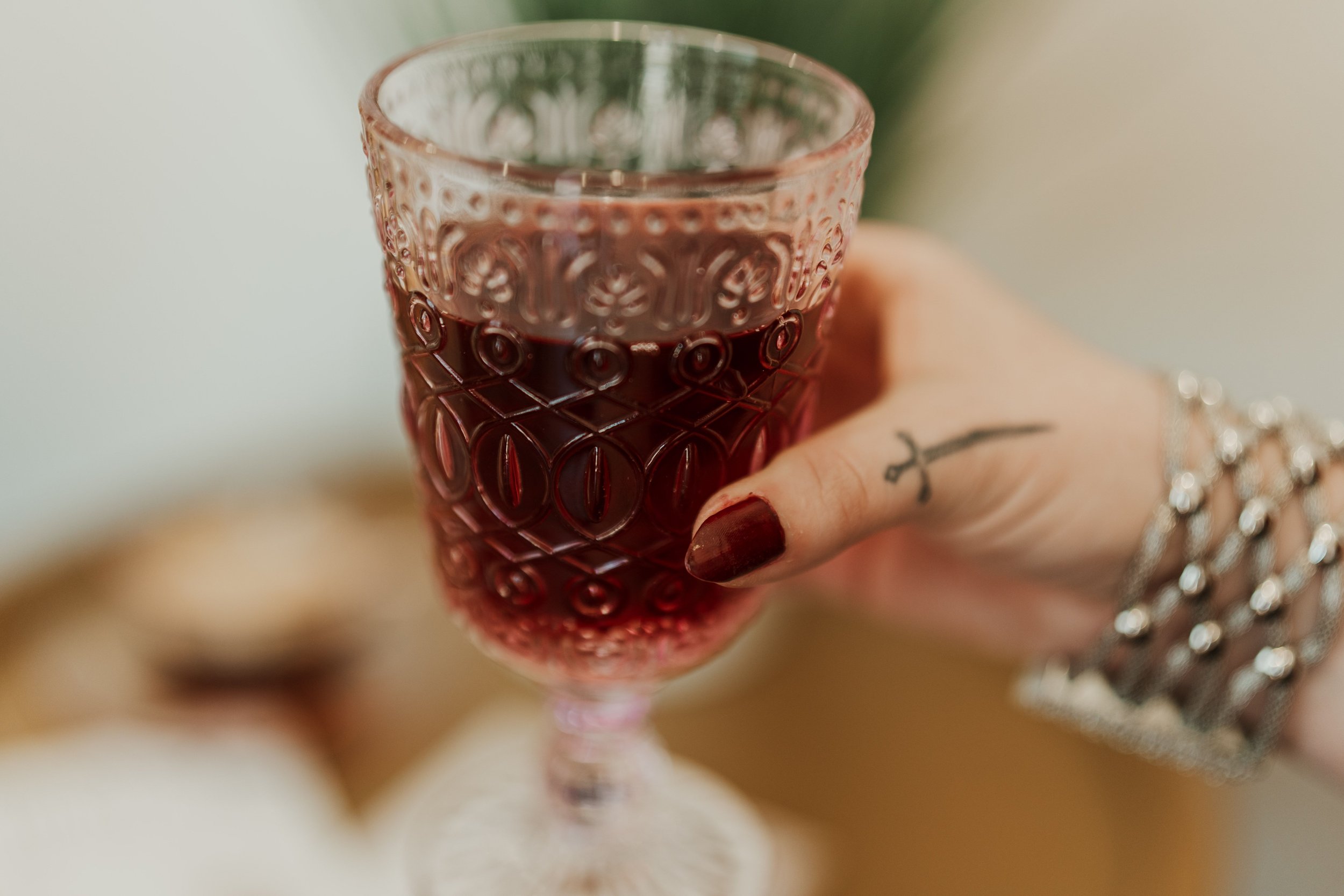 Close-up of a hand holding a glass of red wine with detailed embossed patterns, some rings on fingers, and a tattoo visible on the index finger.
