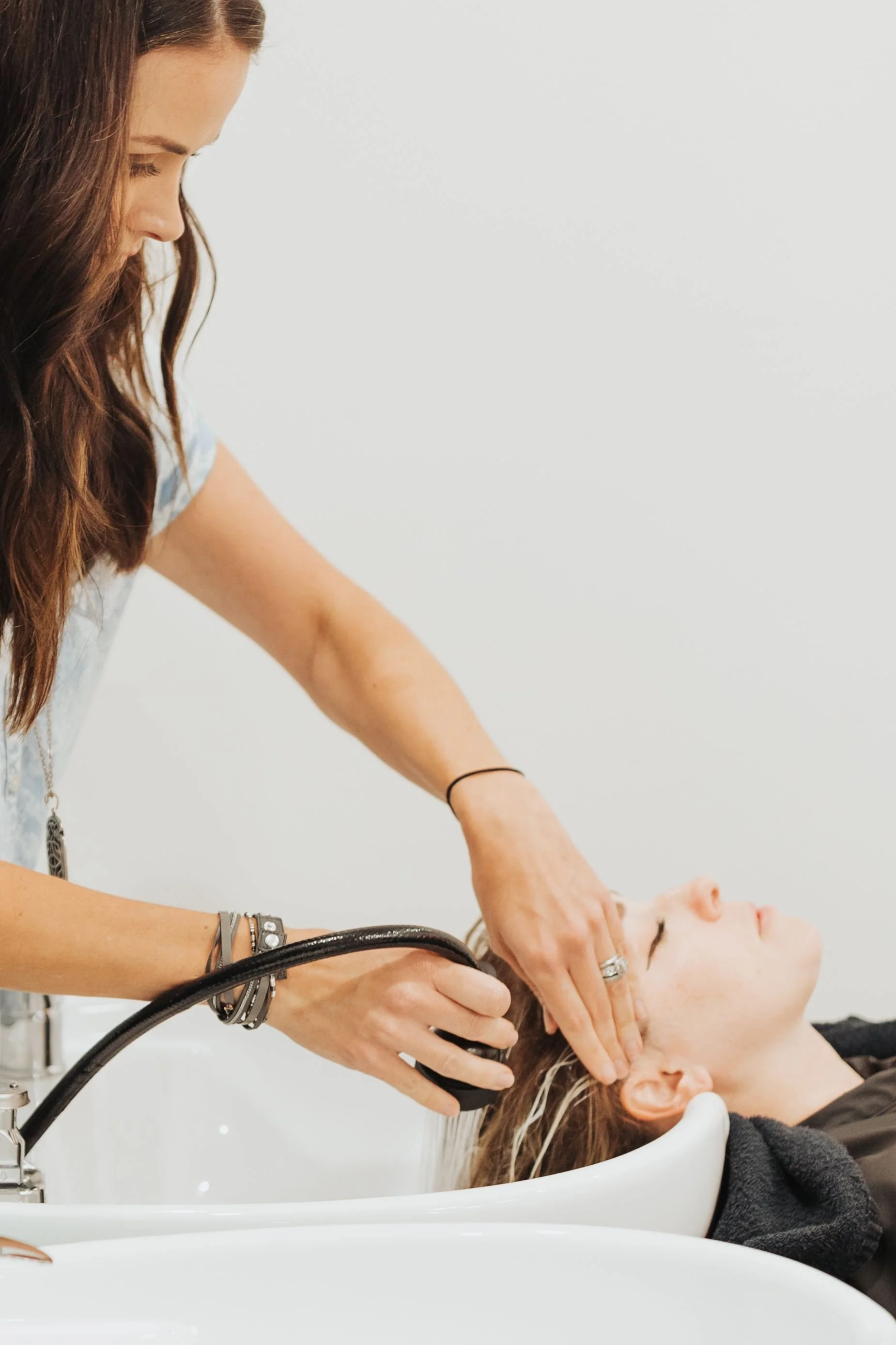 A woman with long brown hair getting her hair washed at a salon while a stylist rinses her hair over a white sink.