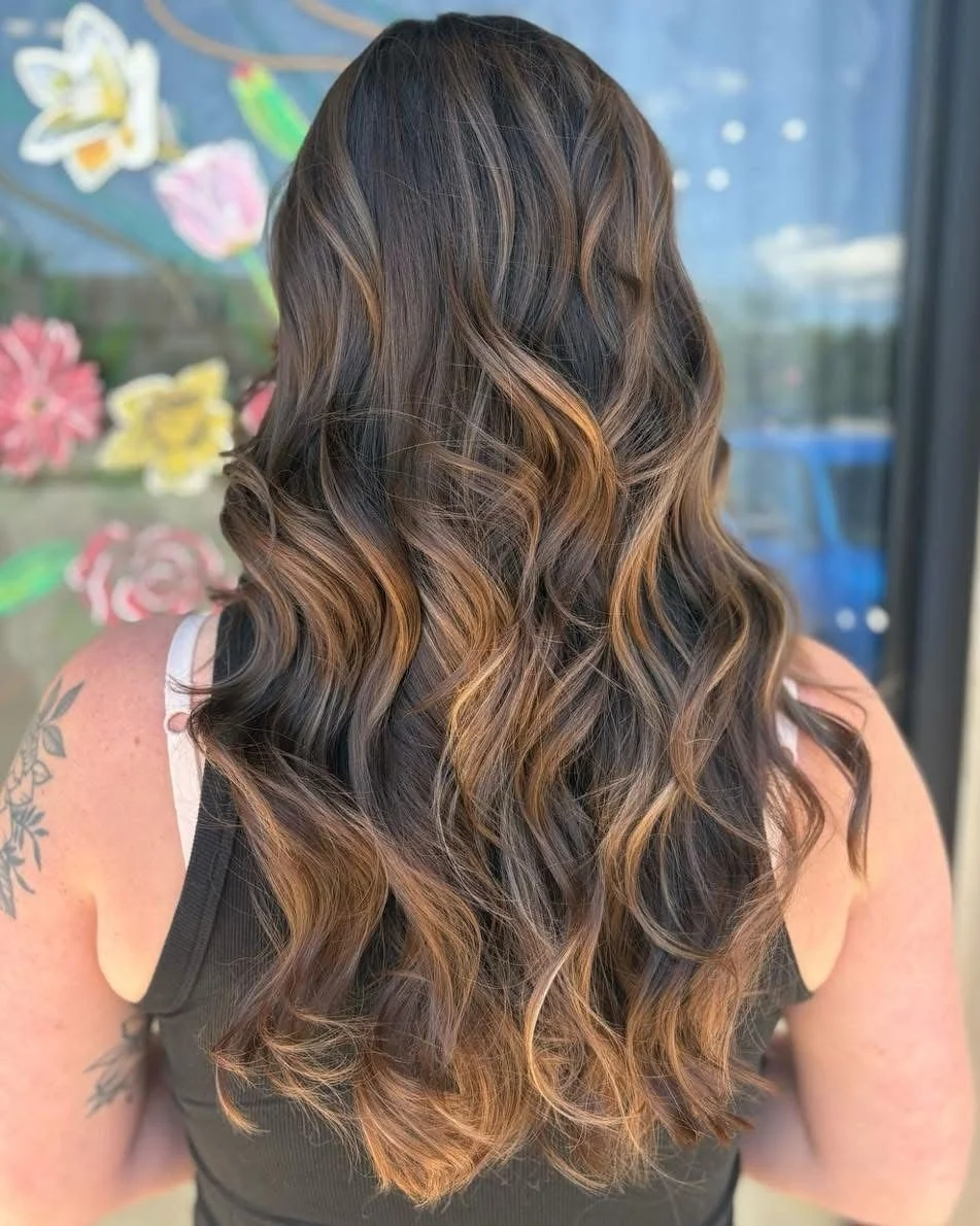 Back view of a woman with long, wavy brown hair with caramel highlights, wearing a black sleeveless top, standing in front of a window with colorful butterfly and flower decorations.