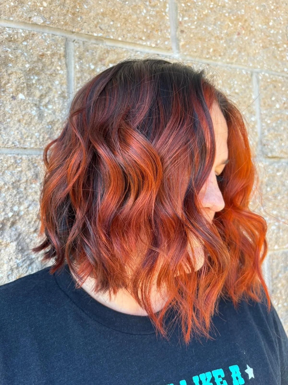 A woman with shoulder-length wavy hair dyed in shades of red and orange, standing against a beige brick wall.