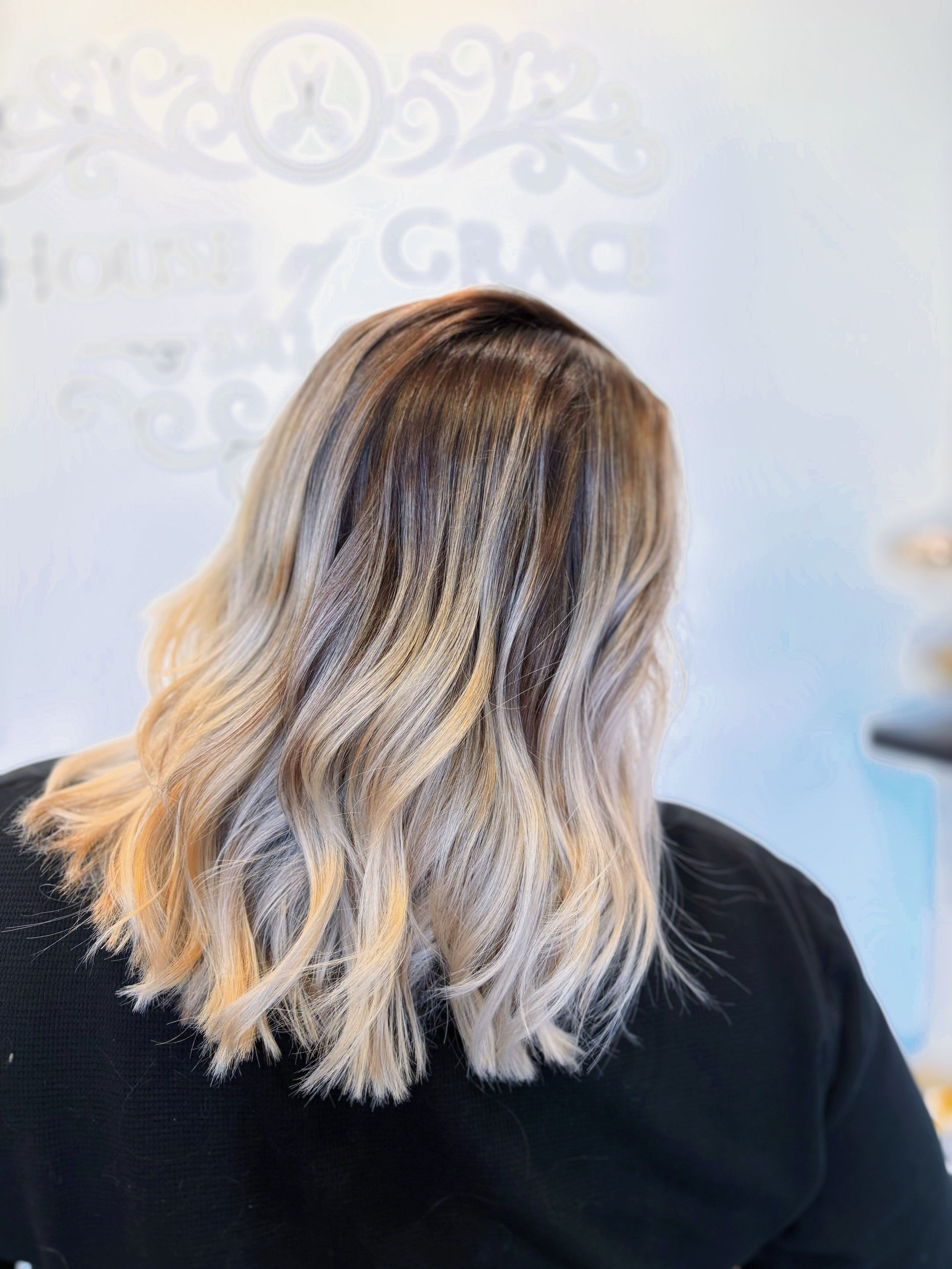 Woman with long, wavy blonde hair sitting in a salon, facing away from the camera.