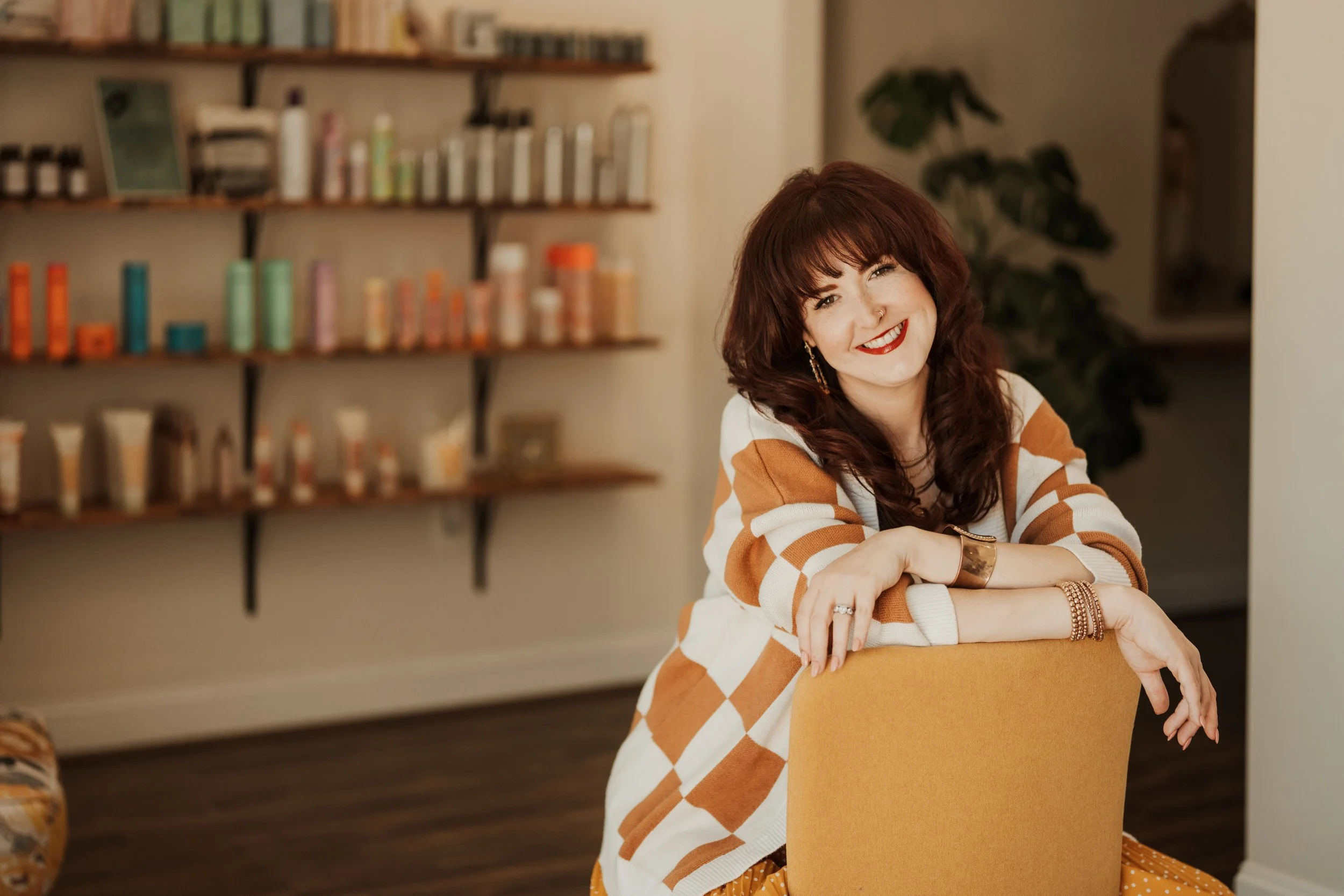A woman with red hair, wearing a white and orange argyle patterned sweater, smiling, sitting at a table with a yellow folder in front of her, in a cozy room with wooden shelves and books behind her.