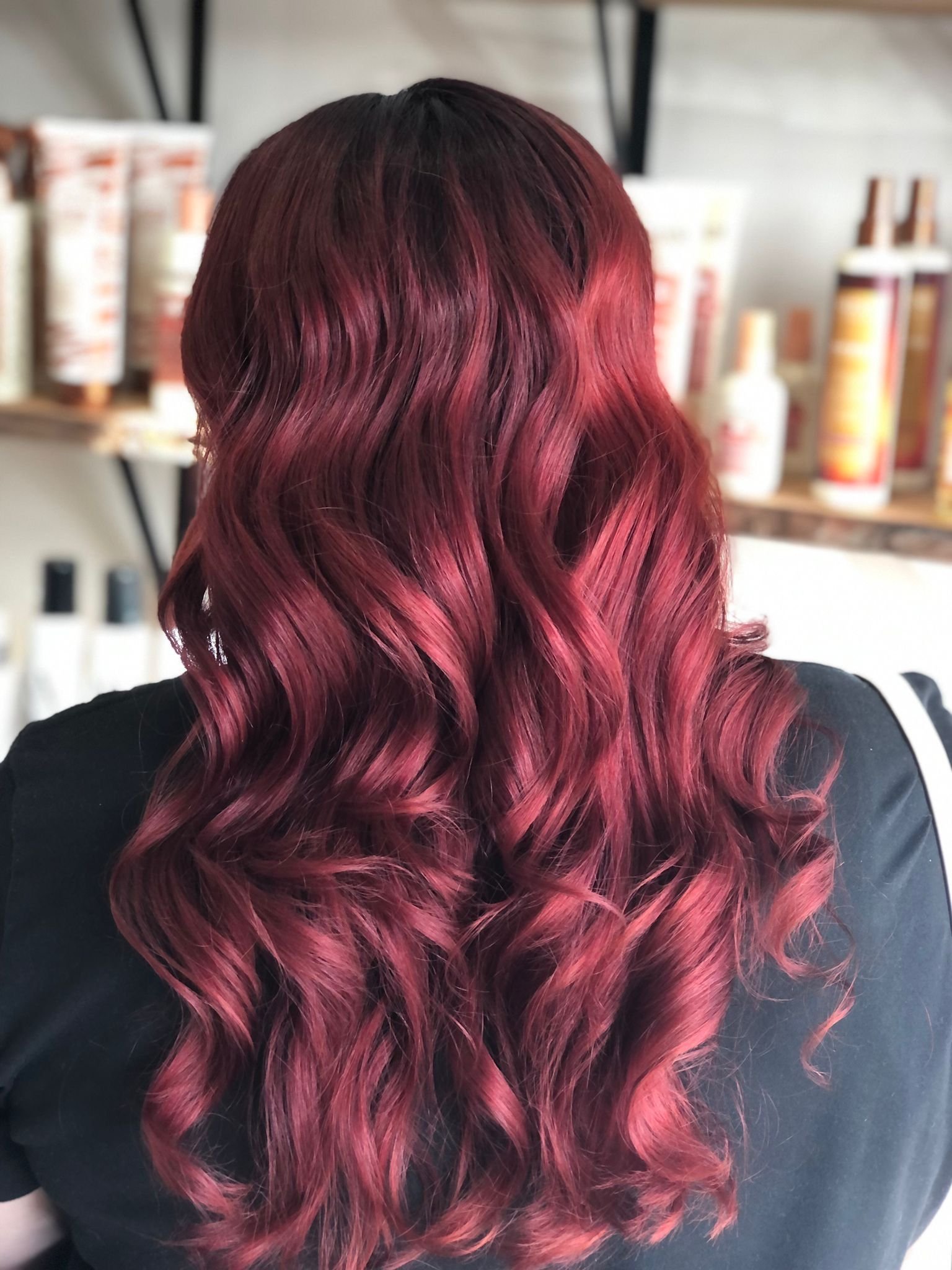 Close-up of a person's wavy hair dyed in shades of burgundy and red, with a darker root, styled in loose waves, seen from the back.