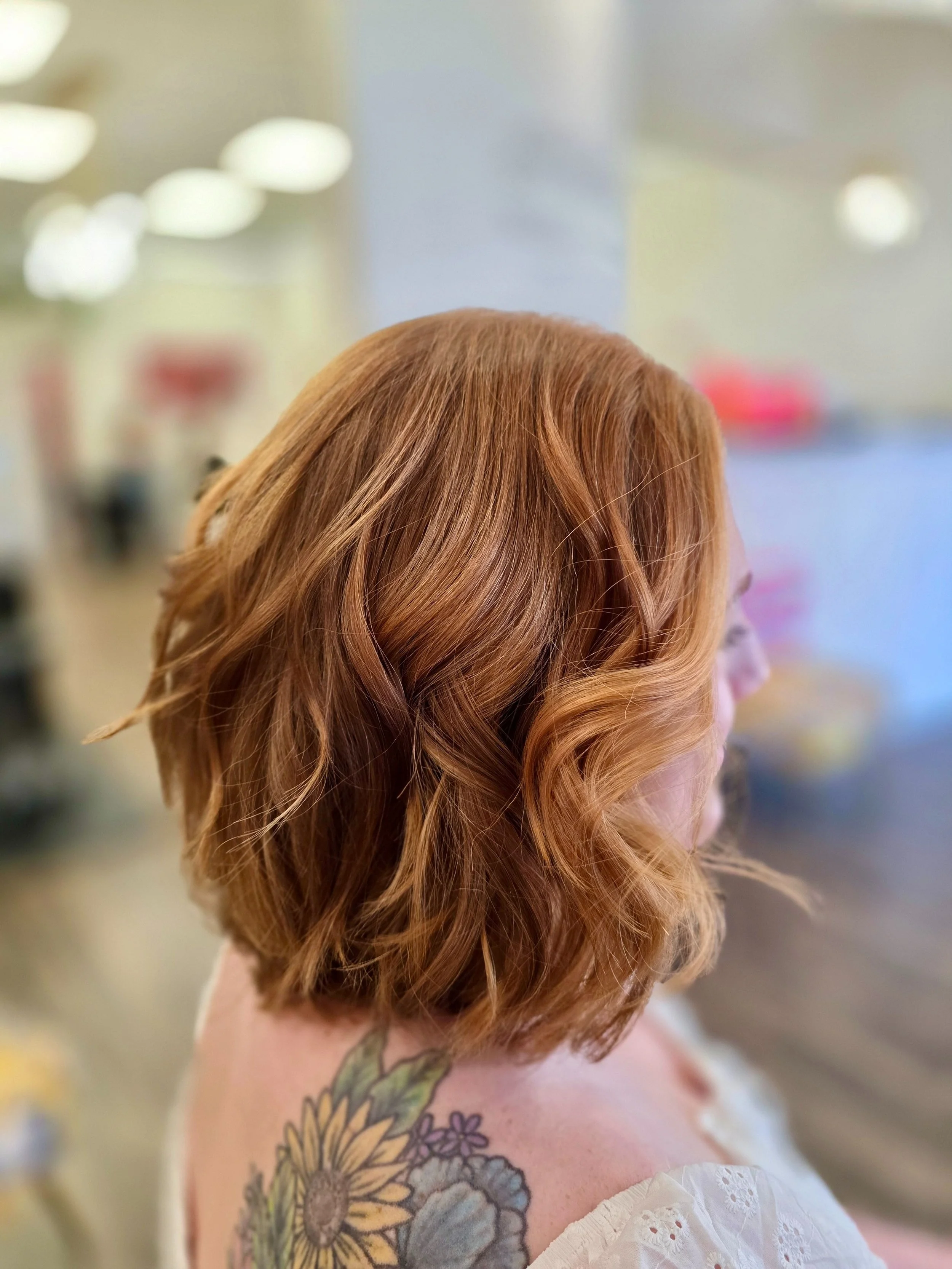 Close-up of a woman with red, wavy hair and a sunflower tattoo on her shoulder, wearing a white off-shoulder top.