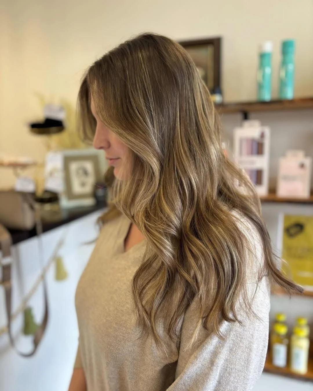 Side view of a woman with wavy, long brown hair wearing a beige sweater in a salon background.