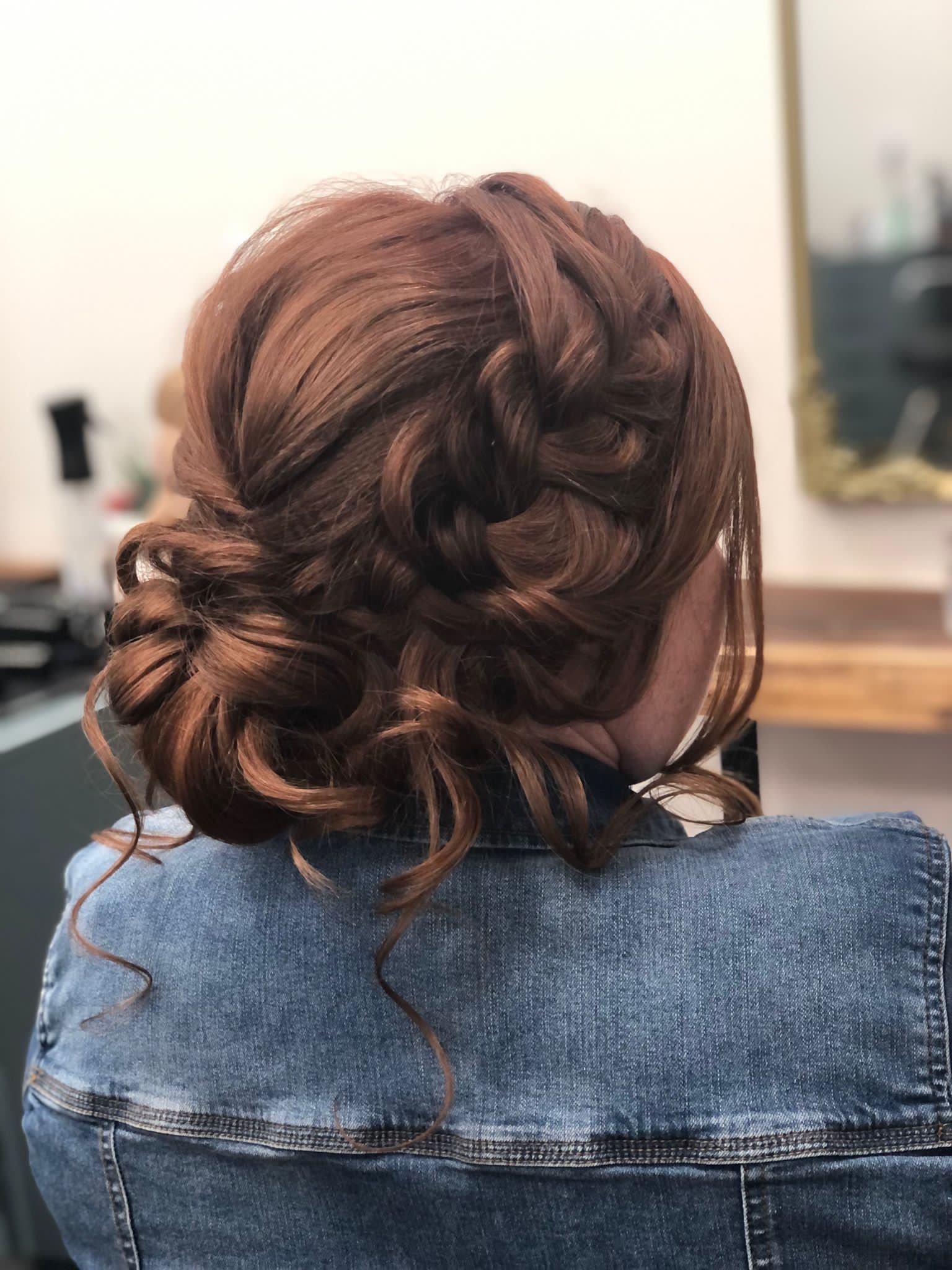 Woman with styled, curly, auburn hair, wearing a denim jacket, sitting with her back to the camera in a salon or similar setting.