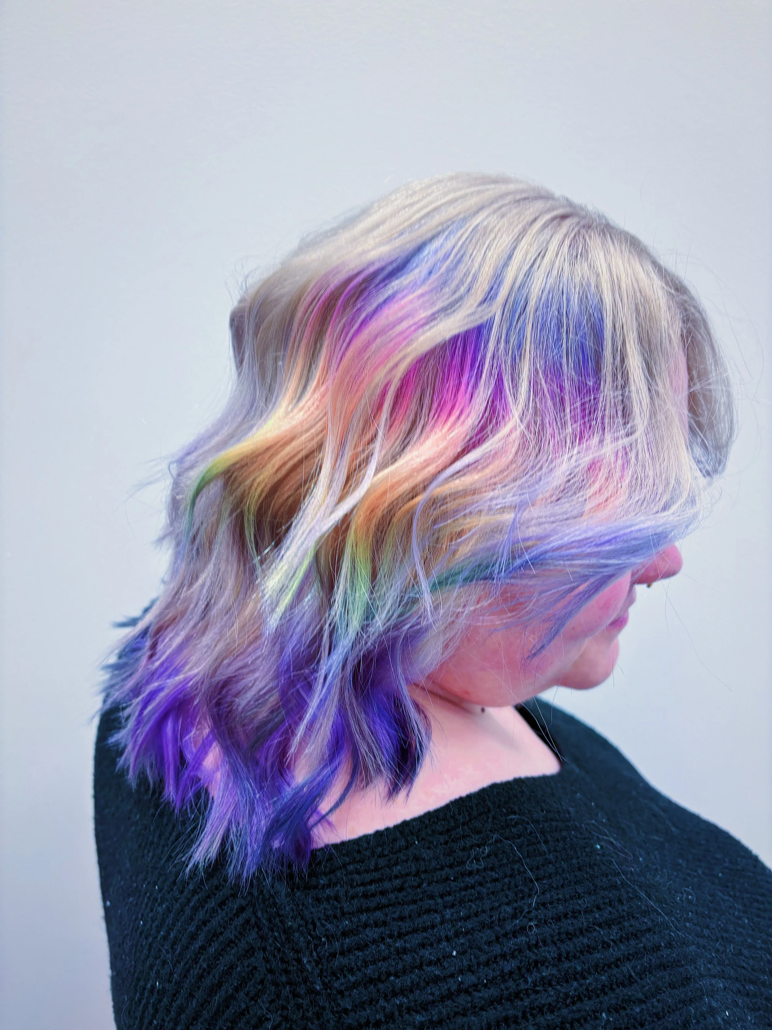 Side view of a woman with flowing rainbow-colored hair, wearing a black textured top, against a plain light background.