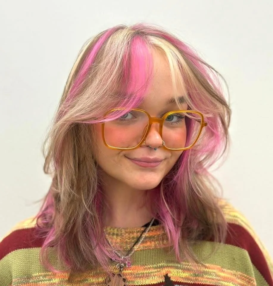 Young woman with pink and blonde hair, wearing oversized orange glasses, a septum piercing, and a colorful striped sweater.