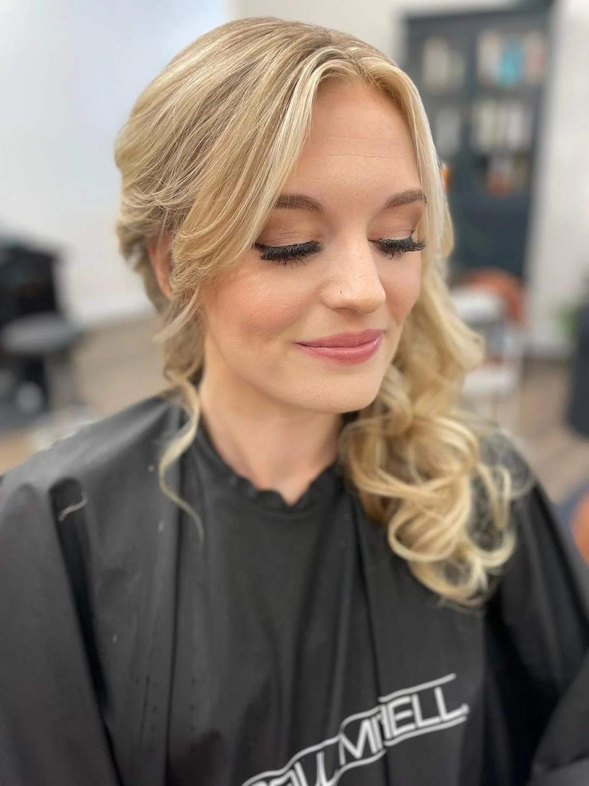A woman with blonde, wavy hair and closed eyes, smiling softly, in a salon chair wearing a black salon cape.