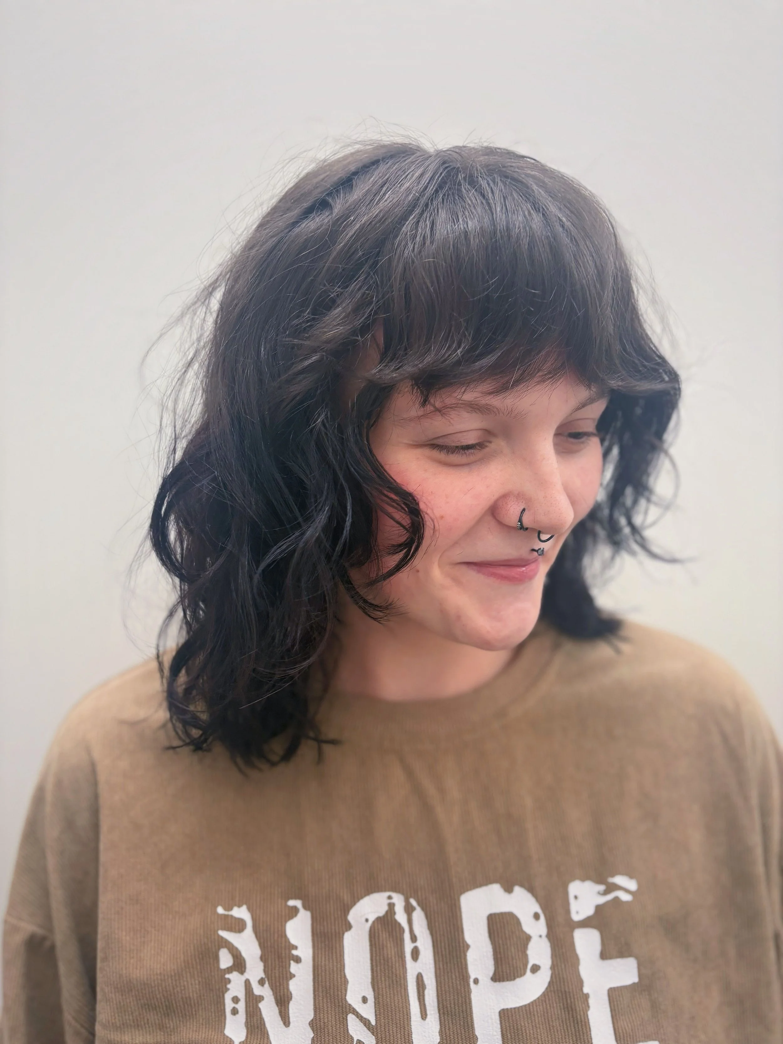 A young woman with dark, curly hair and multiple facial piercings, including a septum ring and lip rings, standing against a plain, light-colored background, wearing a light brown sweatshirt with white text.
