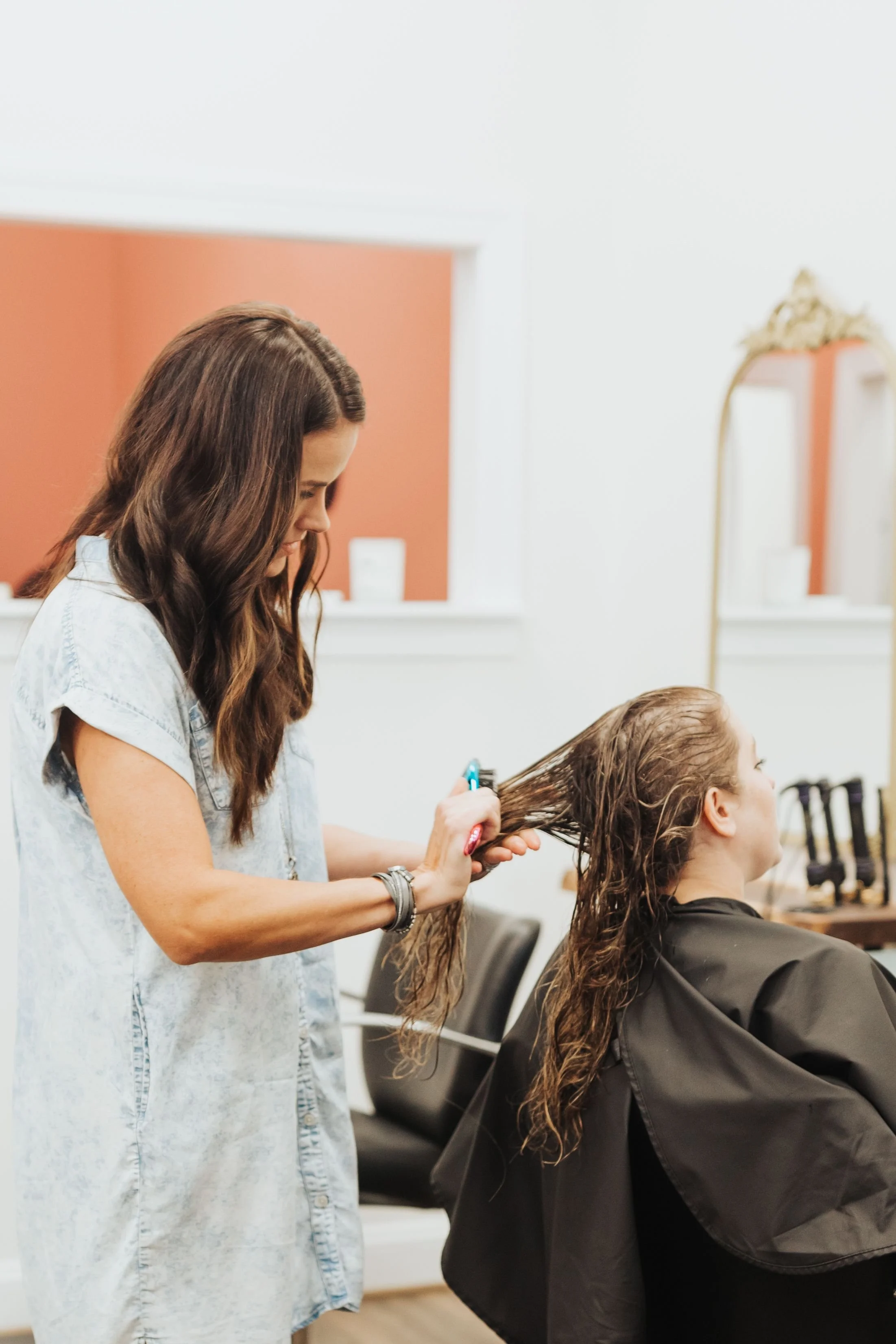 A hairstylist with wavy brown hair and a denim shirt is blow drying and styling a woman's long, brown, curly hair in a salon. The client is seated with a black cape around her shoulders, and there are salon chairs and a mirror with an ornate gold frame on the wall behind them.