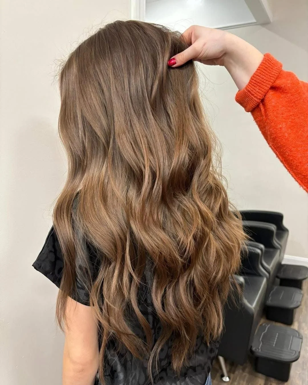 A woman with long wavy light brown hair, wearing a black top, is having her hair styled or examined at a salon, with a person in an orange sleeve holding her hair.