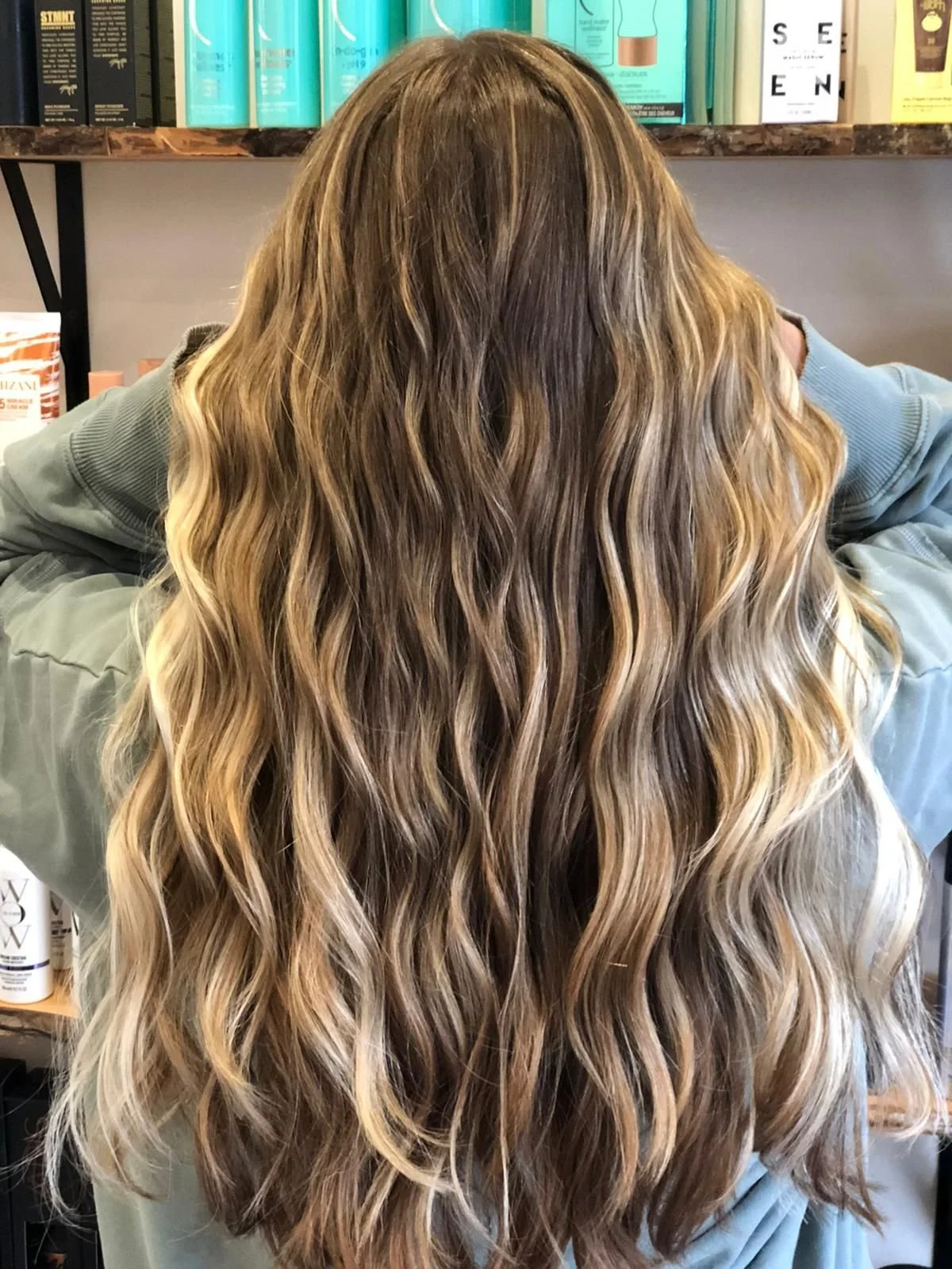 Person with long, wavy, blonde hair sitting in a salon with shelves of hair products in the background.
