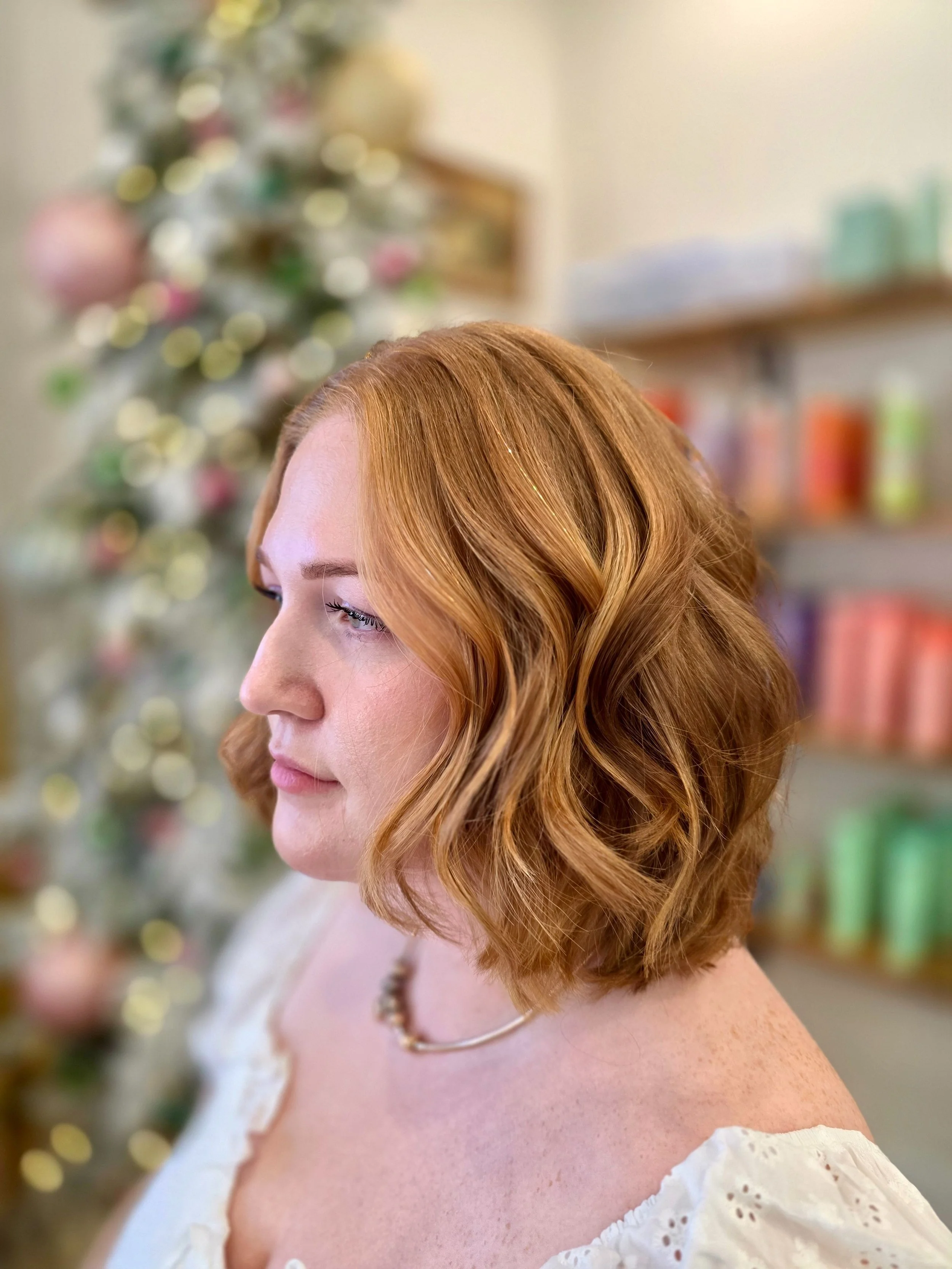 A woman with red hair styled in loose waves, wearing an off-the-shoulder white dress with eyelet details, a pearl necklace, and a gold chain necklace. She is standing indoors with a blurred Christmas tree with ornaments and a bookshelf in the backgro