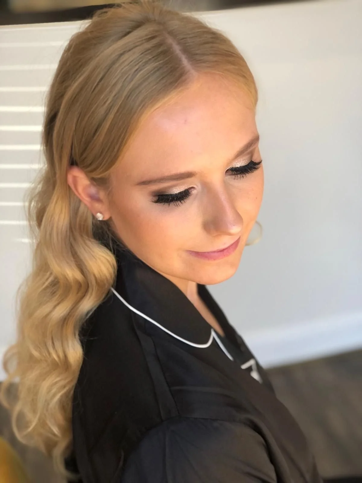 A woman with wavy blonde hair styled in loose curls, wearing makeup with dark eyeliner and mascara, silver stud earrings, and a black outfit with white piping, standing near a white wall with a vent or horizontal slats.