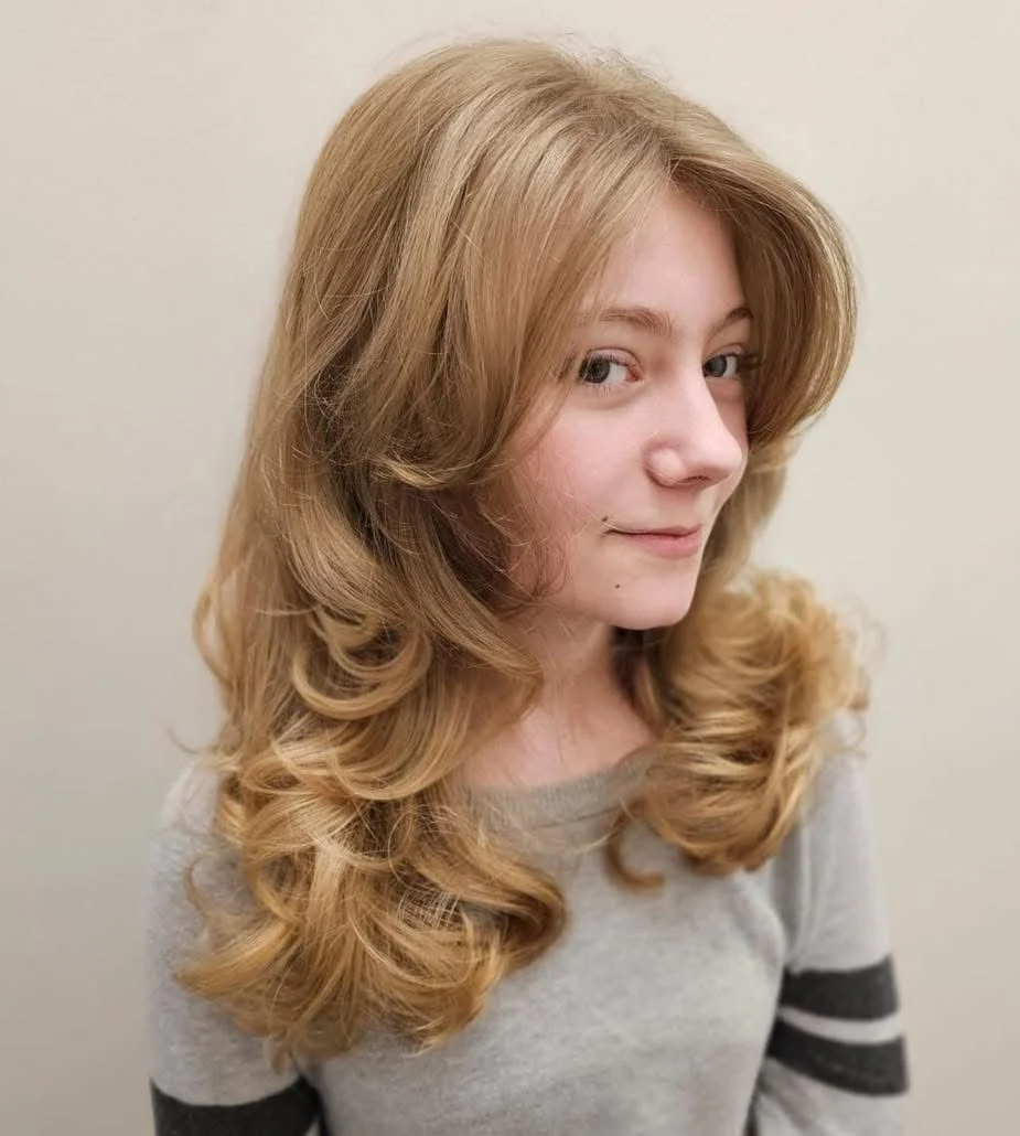 A young woman with long, curly blonde hair, slightly turned to the side, wearing a light gray shirt with dark gray stripes on the sleeves.