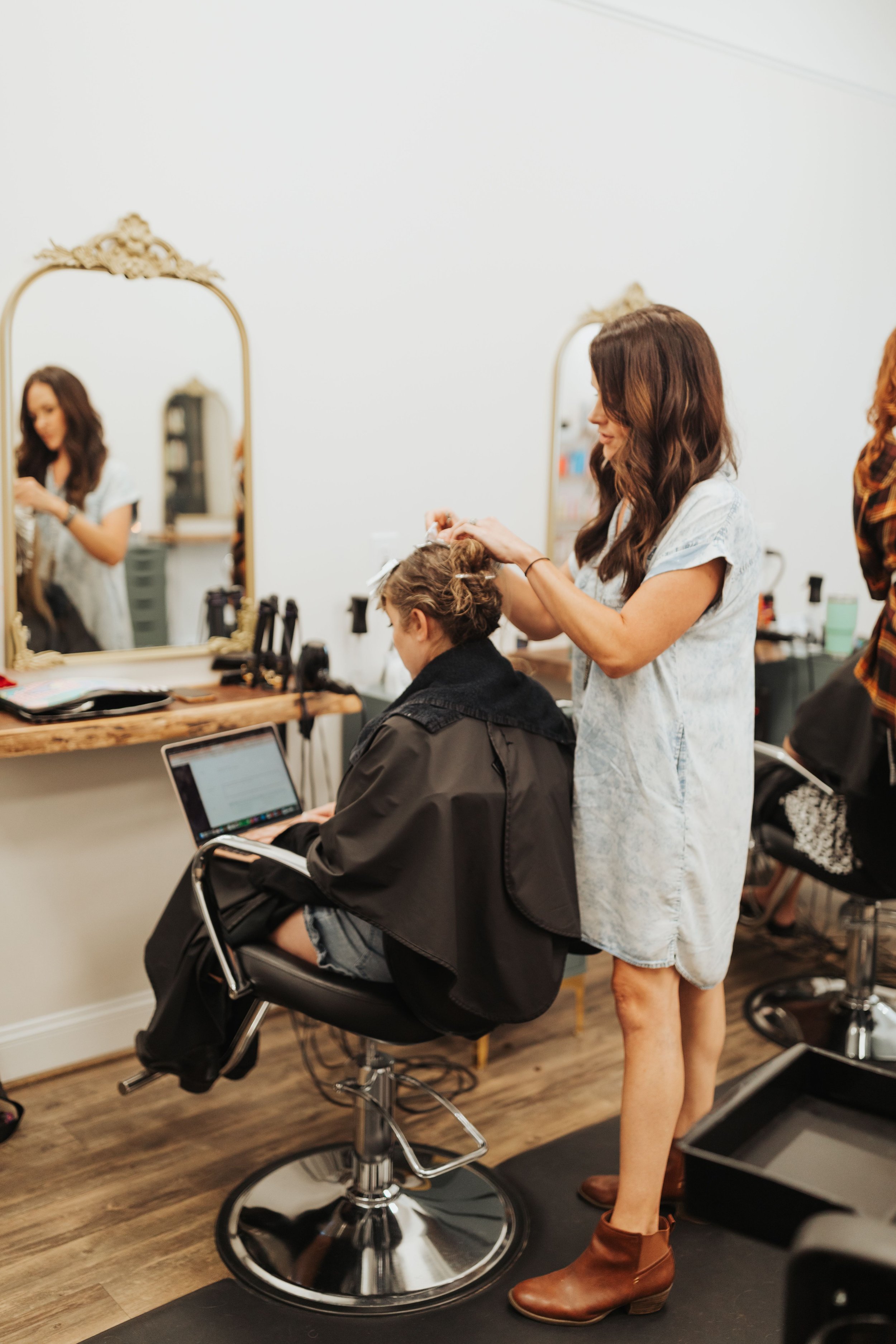 A woman with long brown hair styling a seated woman with curly hair at a hair salon. The stylist is wearing a light-colored dress and brown ankle boots.