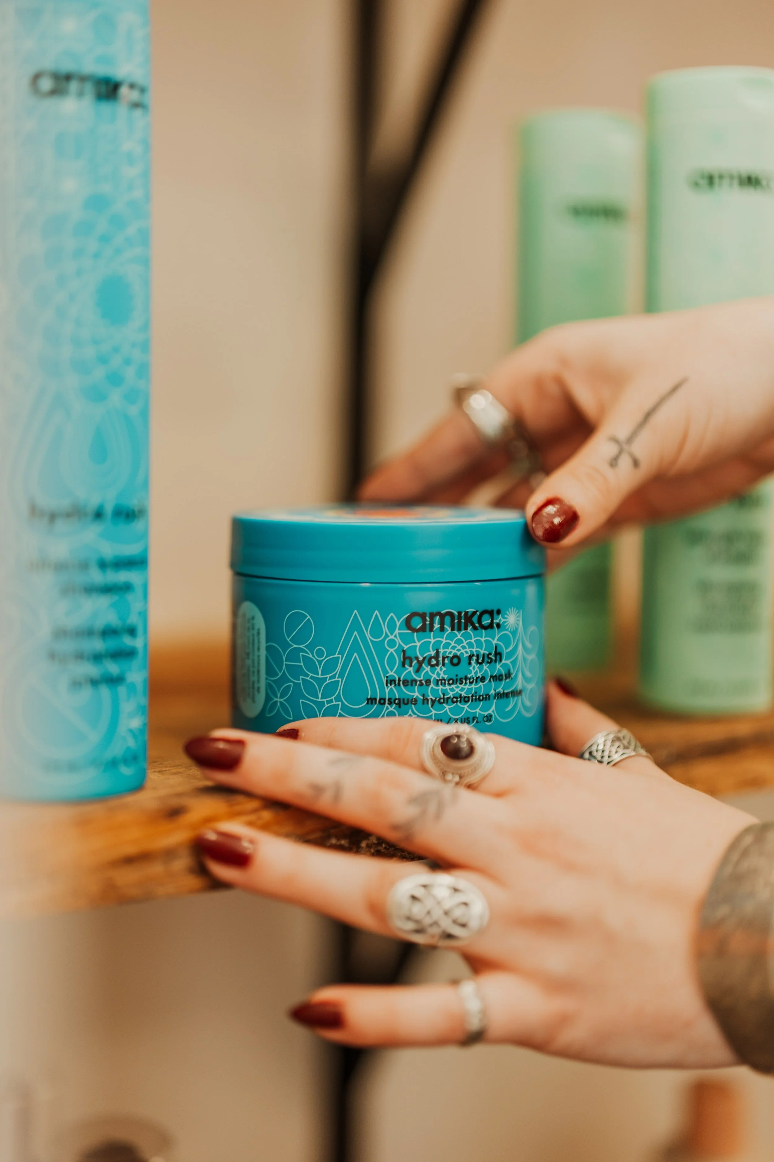 Close-up of a person holding a blue container of Amika Hydro Rush intense moisture mask, with their fingernails painted dark red and multiple silver rings on their fingers. In the background, there are other hair care products and a blurred interior setting.