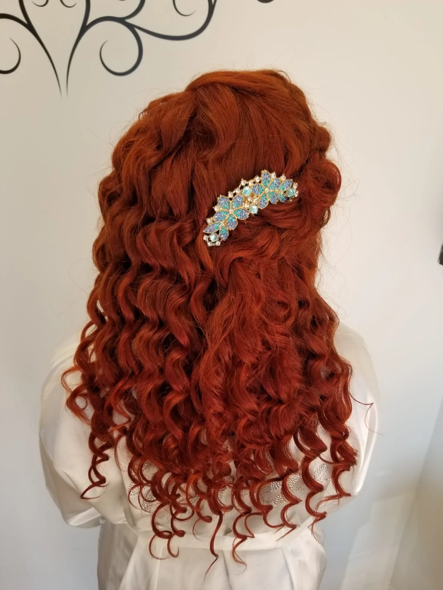 Back view of a woman with long, curly, red hair styled with a decorative hair clip, wearing a white outfit, standing against a plain wall.