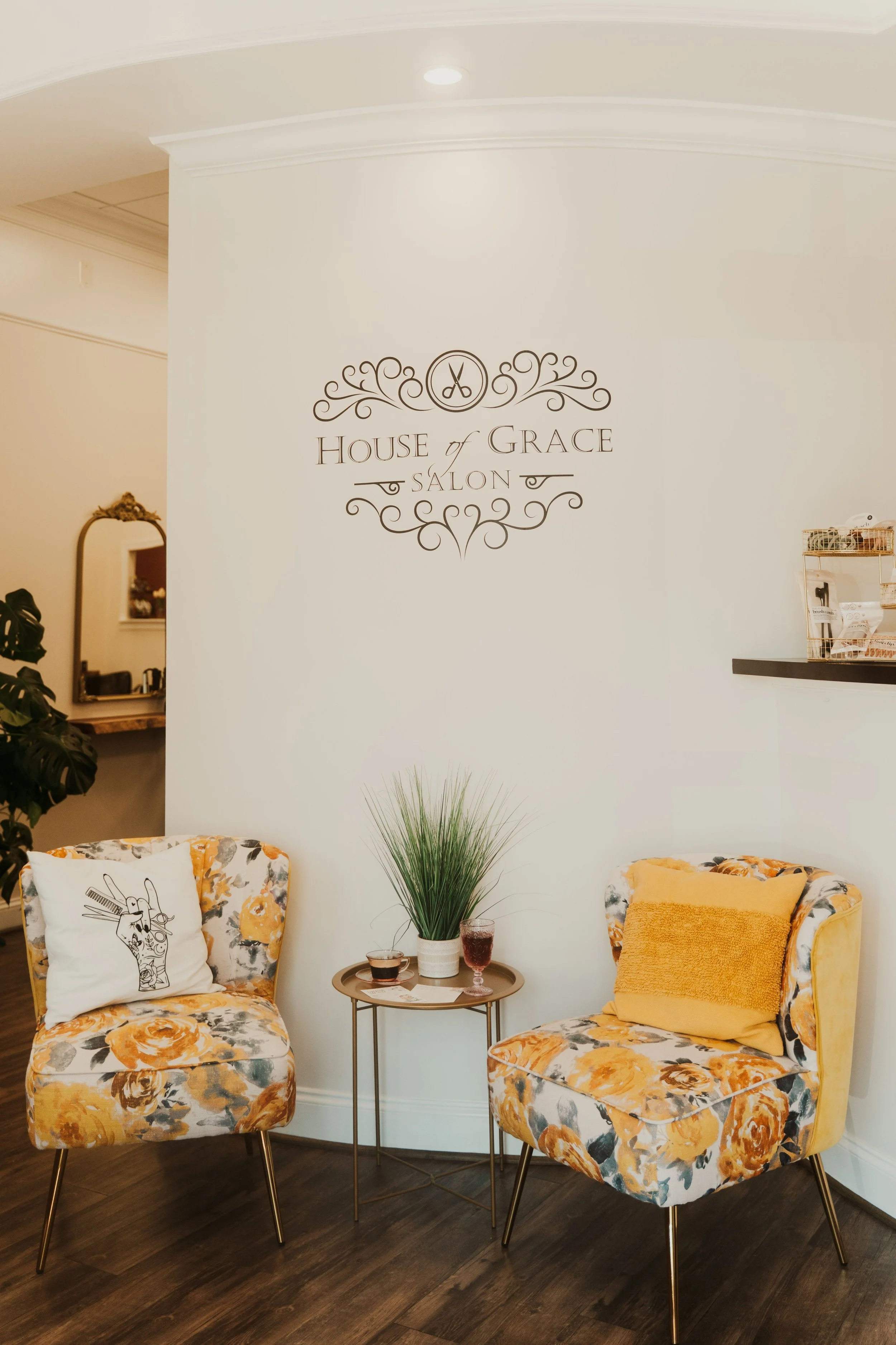 Two floral-patterned chairs with yellow, grey, and white flowers, each with a pillow, one pillow is white with a hairdresser scissors design, the other is yellow. A small round side table with glasses of wine and a potted plant between the chairs, in a salon or waiting area.