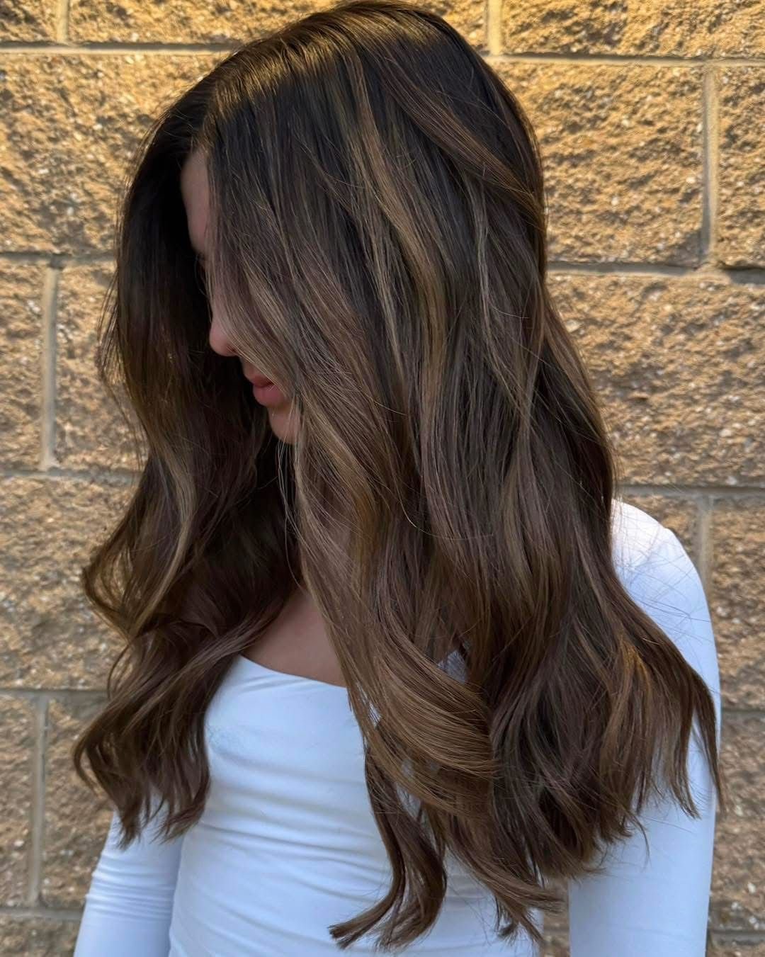 A woman with long, wavy brown hair with blonde highlights, standing against a textured brick wall, looking down.