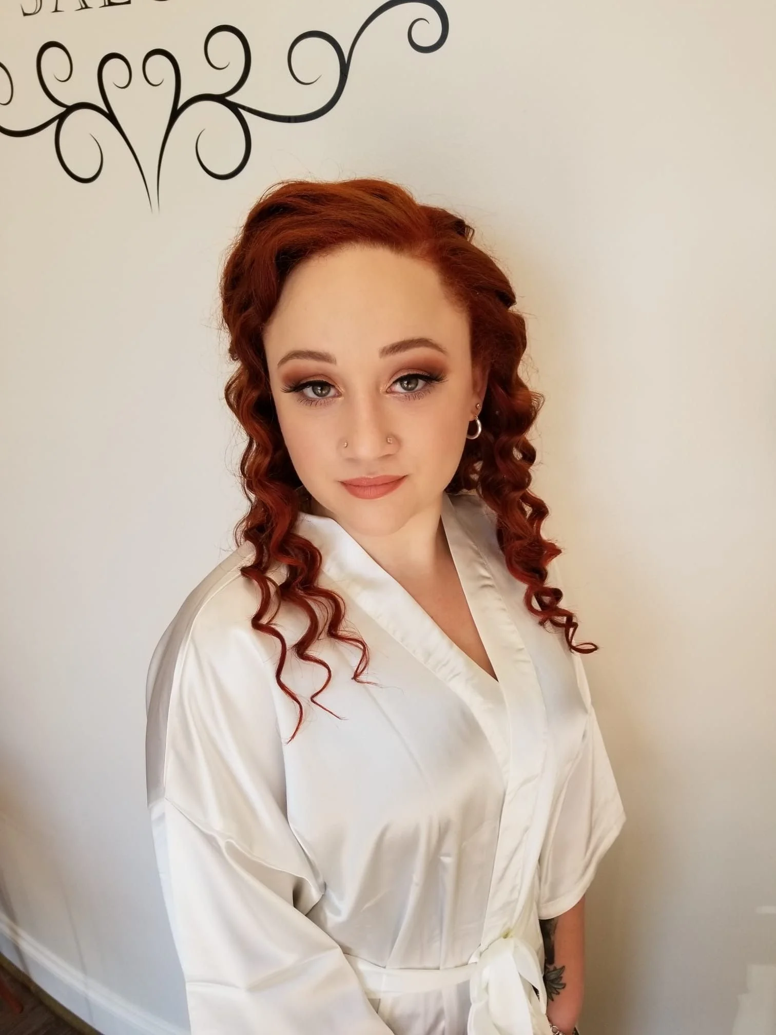 Young woman with red curly hair, wearing makeup and small hoop earrings, dressed in a white satin robe, standing against a white wall with black decorative design.