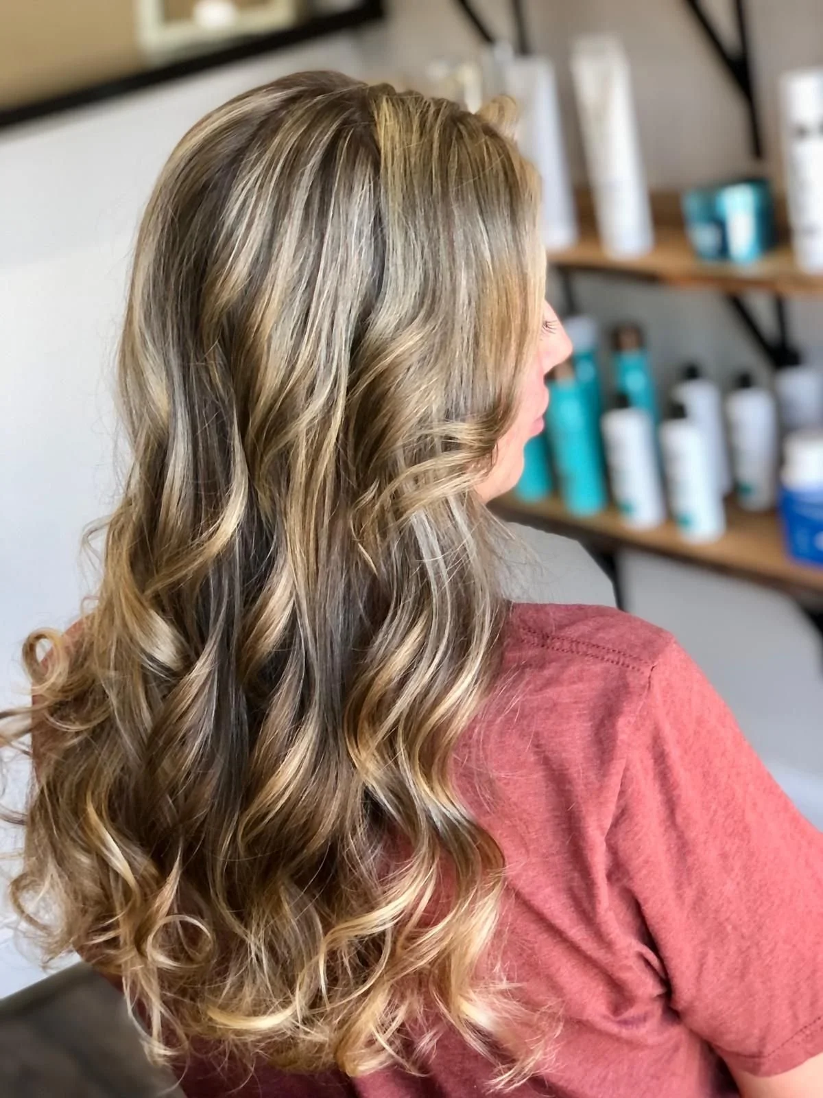 A woman with long, wavy, blonde hair sitting in a salon near hair care products on display.