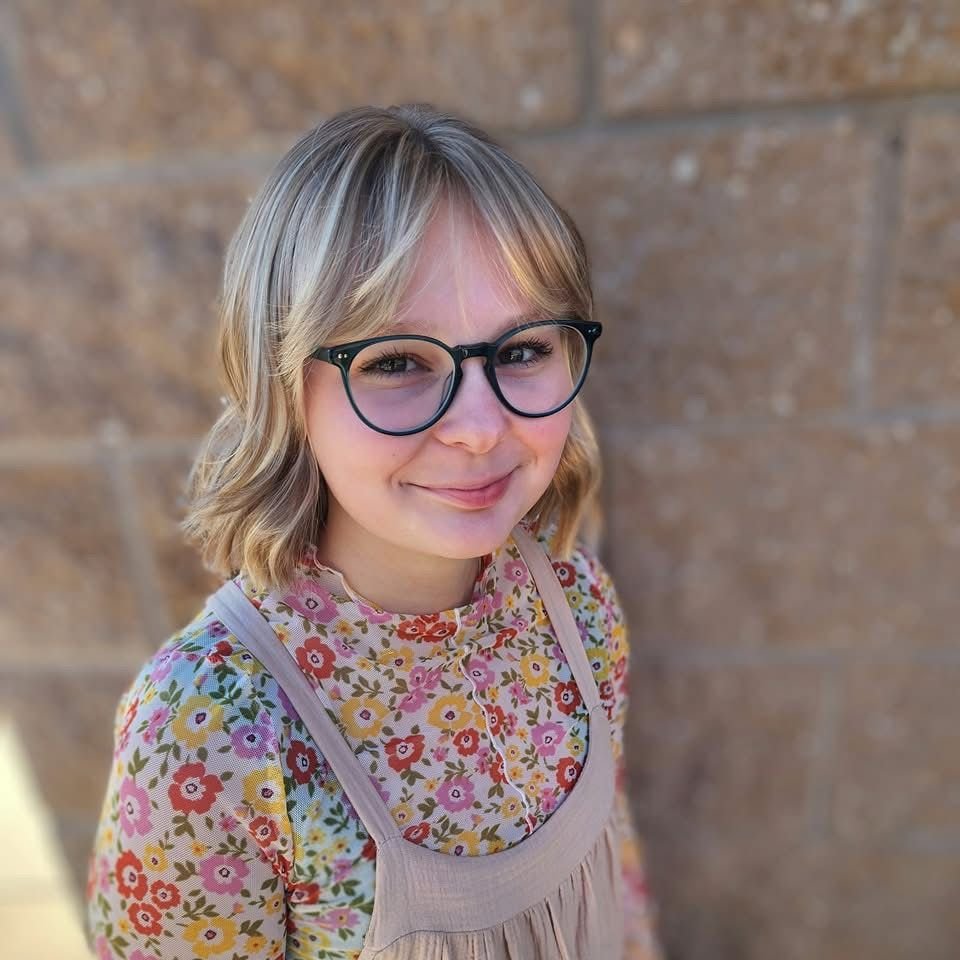 A young girl with blonde hair, glasses, and a floral shirt standing against a brick wall.