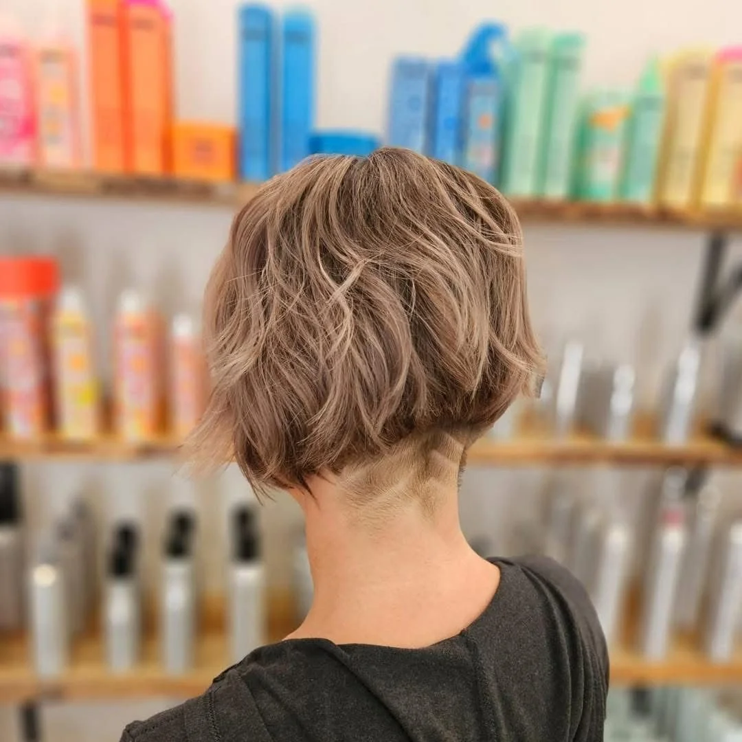 Back view of a woman with a layered short haircut with wavy, ash blonde hair. She is in a store aisle with shelves displaying colorful bottles and boxes.