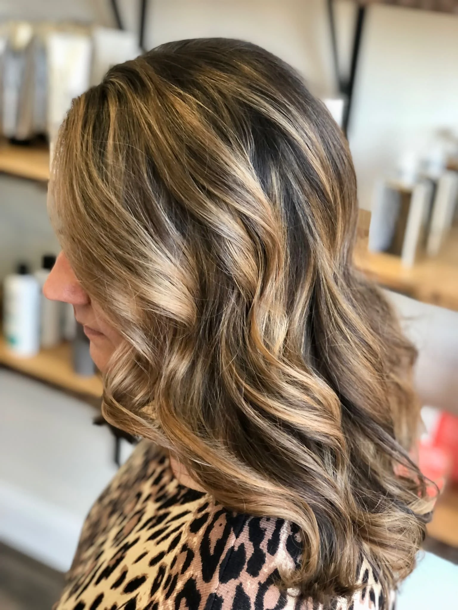 A woman with medium-length, wavy hair featuring highlights of brown and blonde tones, wearing a leopard-print garment, in a salon setting.