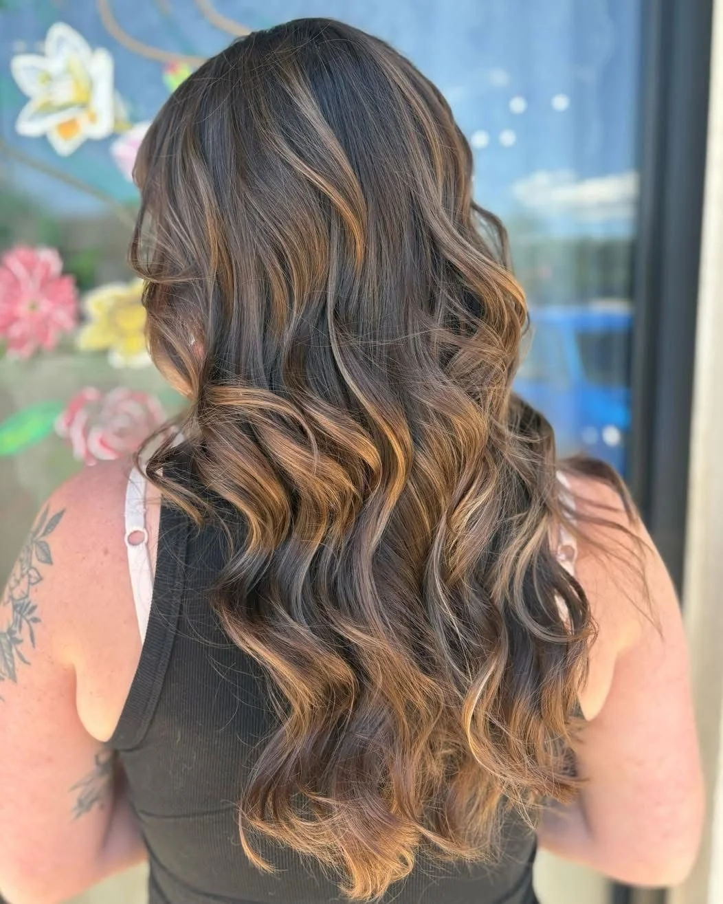 Back view of a woman with long, wavy, brown hair with blonde highlights, wearing a black sleeveless top, standing in front of a colorful floral background.