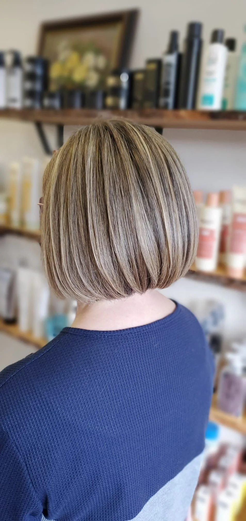 Back view of a woman with a short, straight, blonde bob hairstyle in a salon or store, with shelves of hair products in the background.
