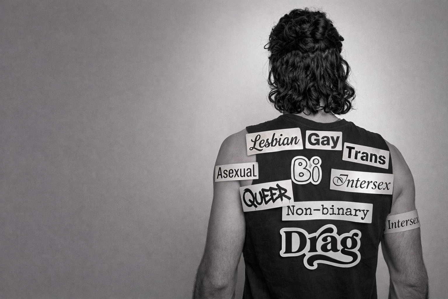 Back view of person with curly hair wearing a sleeveless black shirt with various words and phrases related to LGBTQ+ identities and gender expressions, such as 'Lesbian', 'Gay', 'Trans', 'Asexual', 'Queer', 'Non-binary', 'Intersex', and 'Drag'.