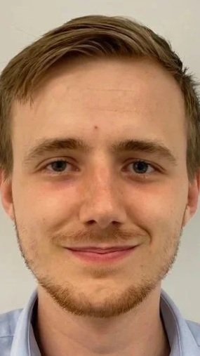 A young man with light brown hair and a slight beard, smiling in front of a plain background.