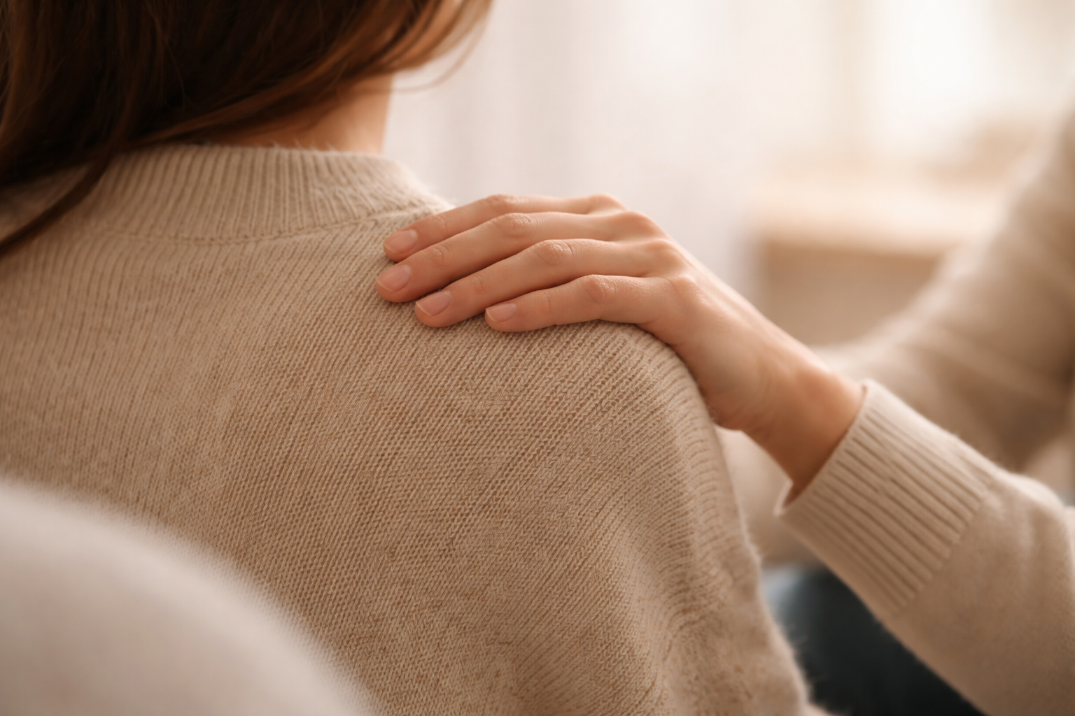 A person with long brown hair wearing a beige sweater, gently placing a hand on their shoulder.