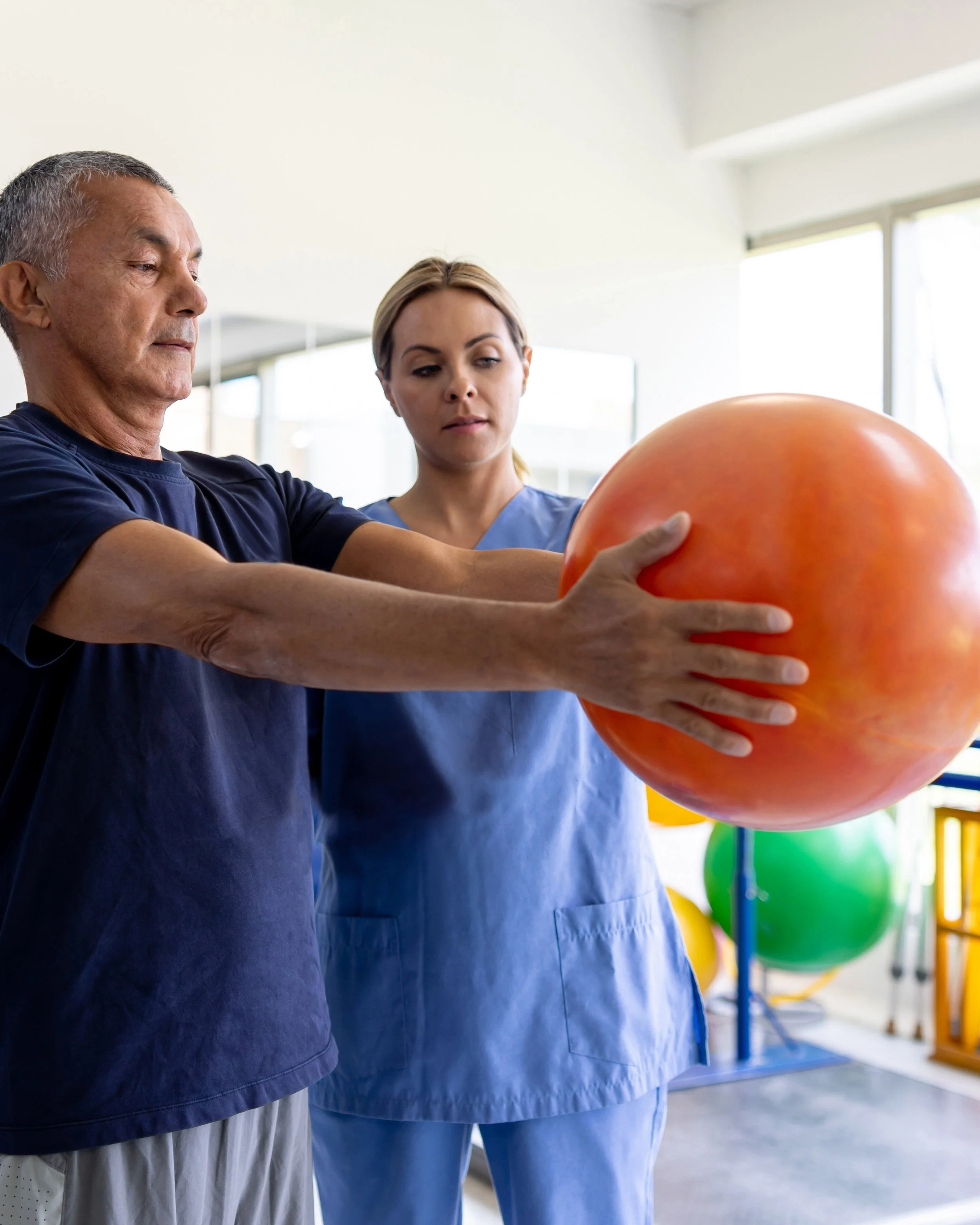 A man and a woman in a fitness or physical therapy facility holding a large orange exercise ball.