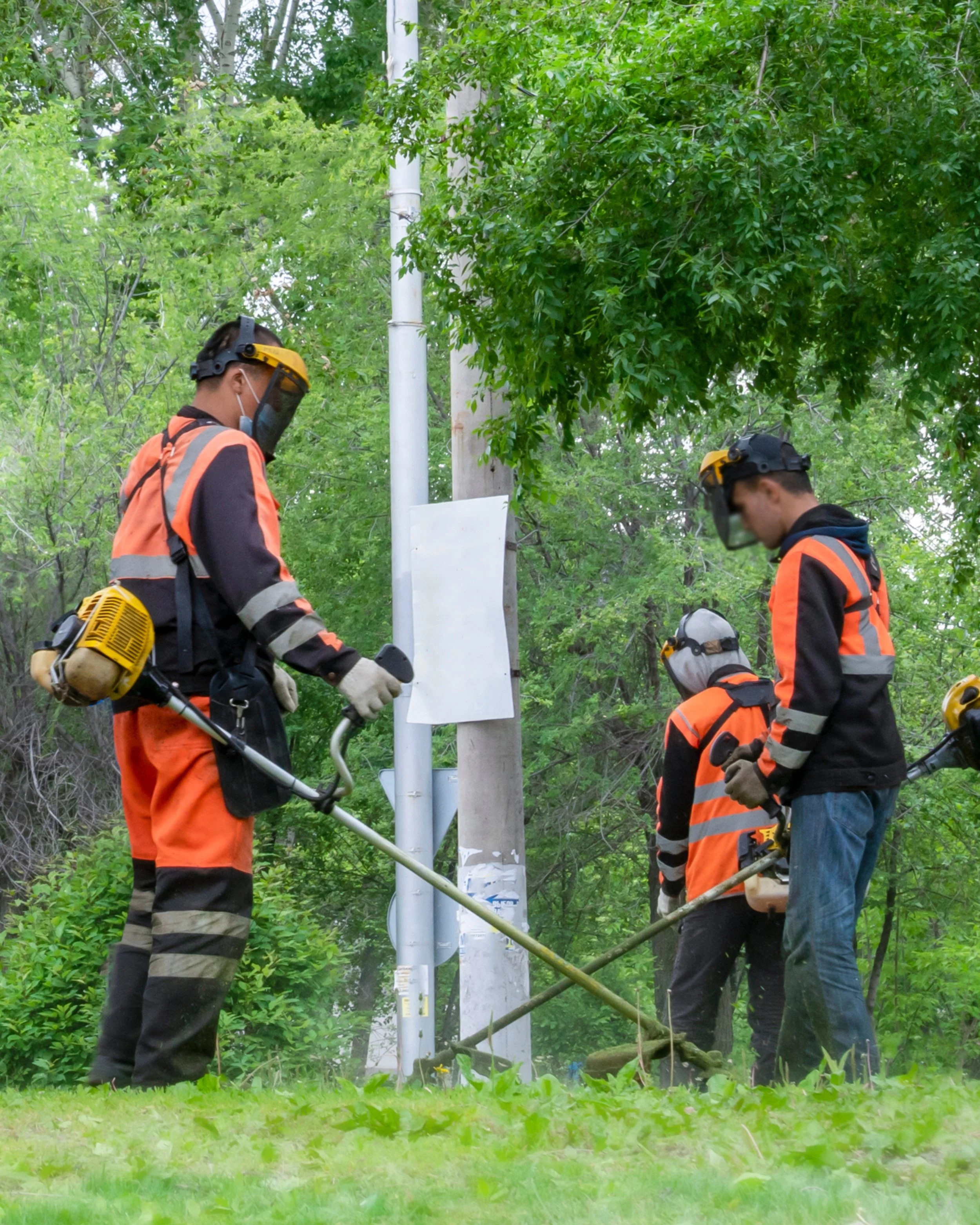 Three workers wearing safety gear using power tools to cut a metal pole in a green outdoor area.