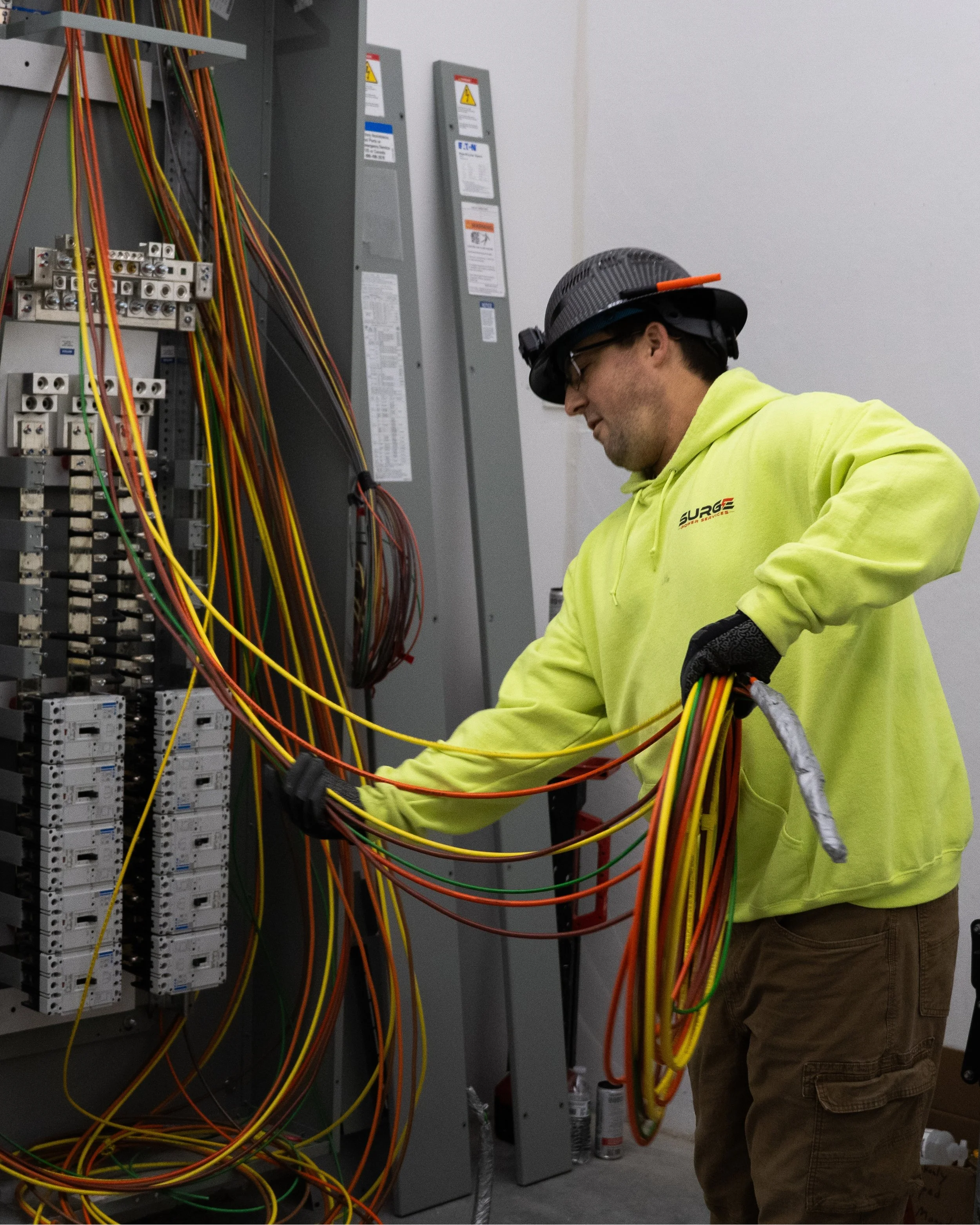 A technician wearing a yellow hoodie, protective gloves, and a safety helmet working with colorful electrical wires inside an electrical panel.