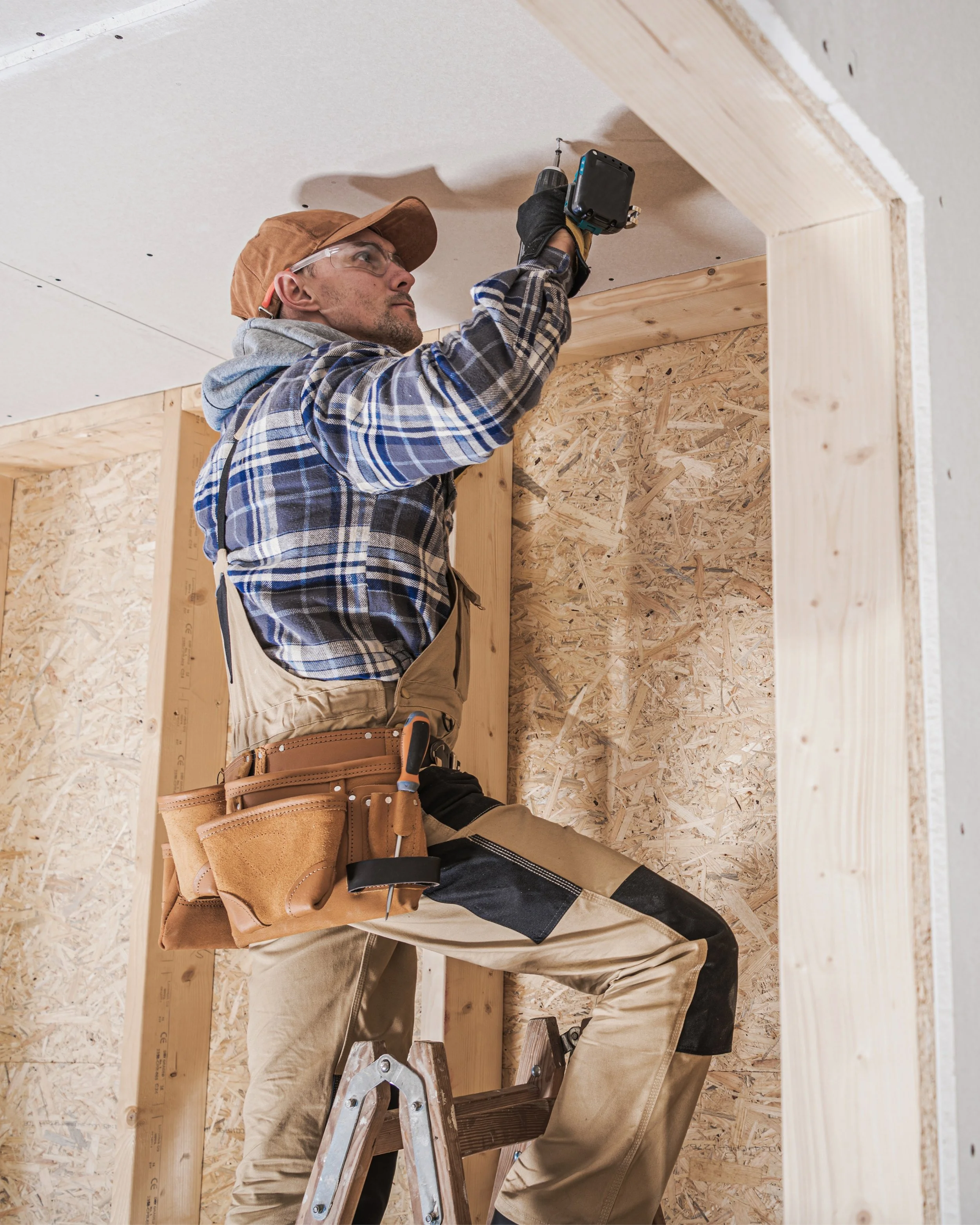 A construction worker wearing a plaid shirt, beige overalls, and a tan cap is using a power drill to work on the ceiling of a room under construction. He is standing on a step ladder, and there are wooden beams and rough OSB wall panels in the background.
