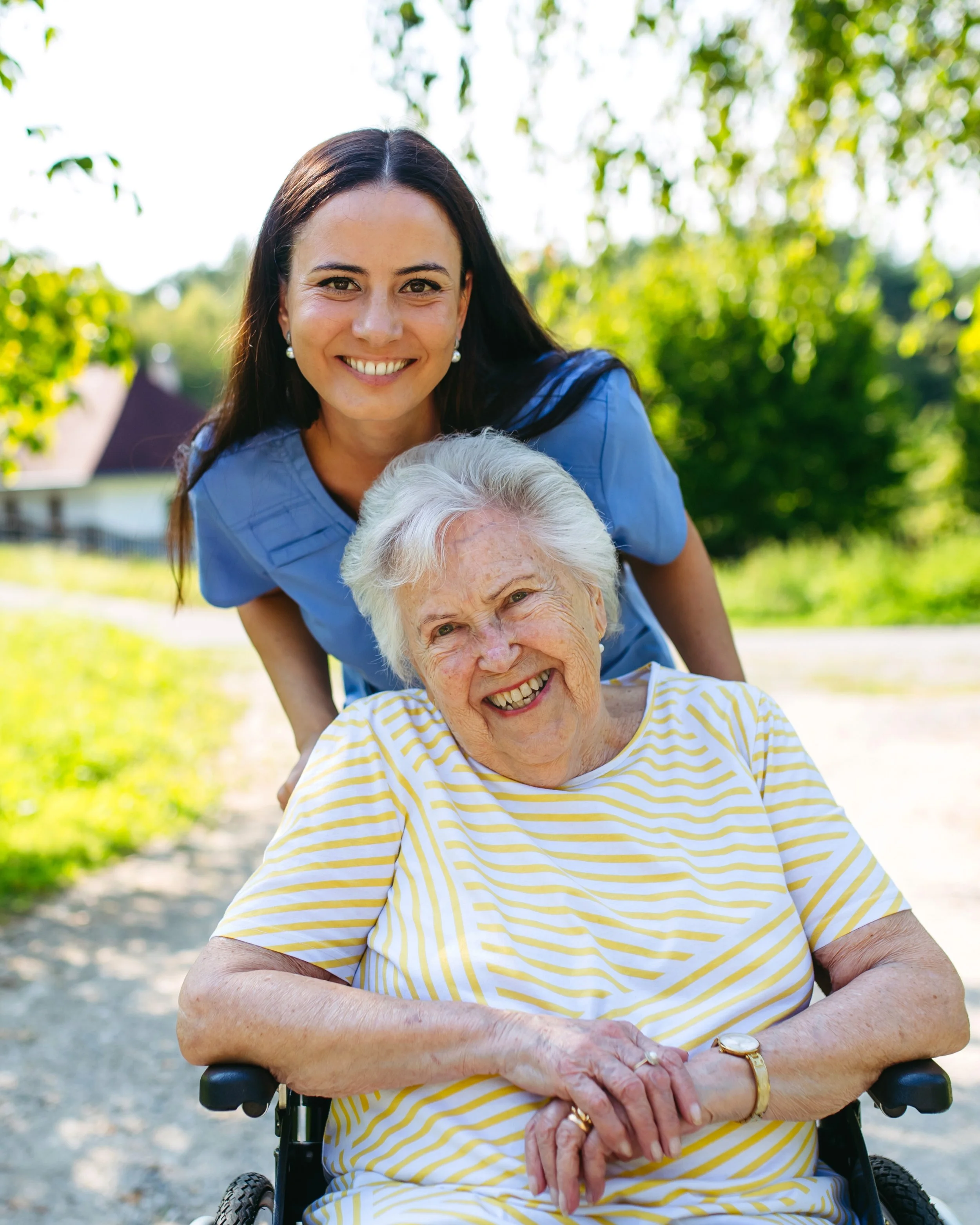 A young female caregiver in a blue uniform smiling behind an elderly woman in a wheelchair outdoors on a sunny day.