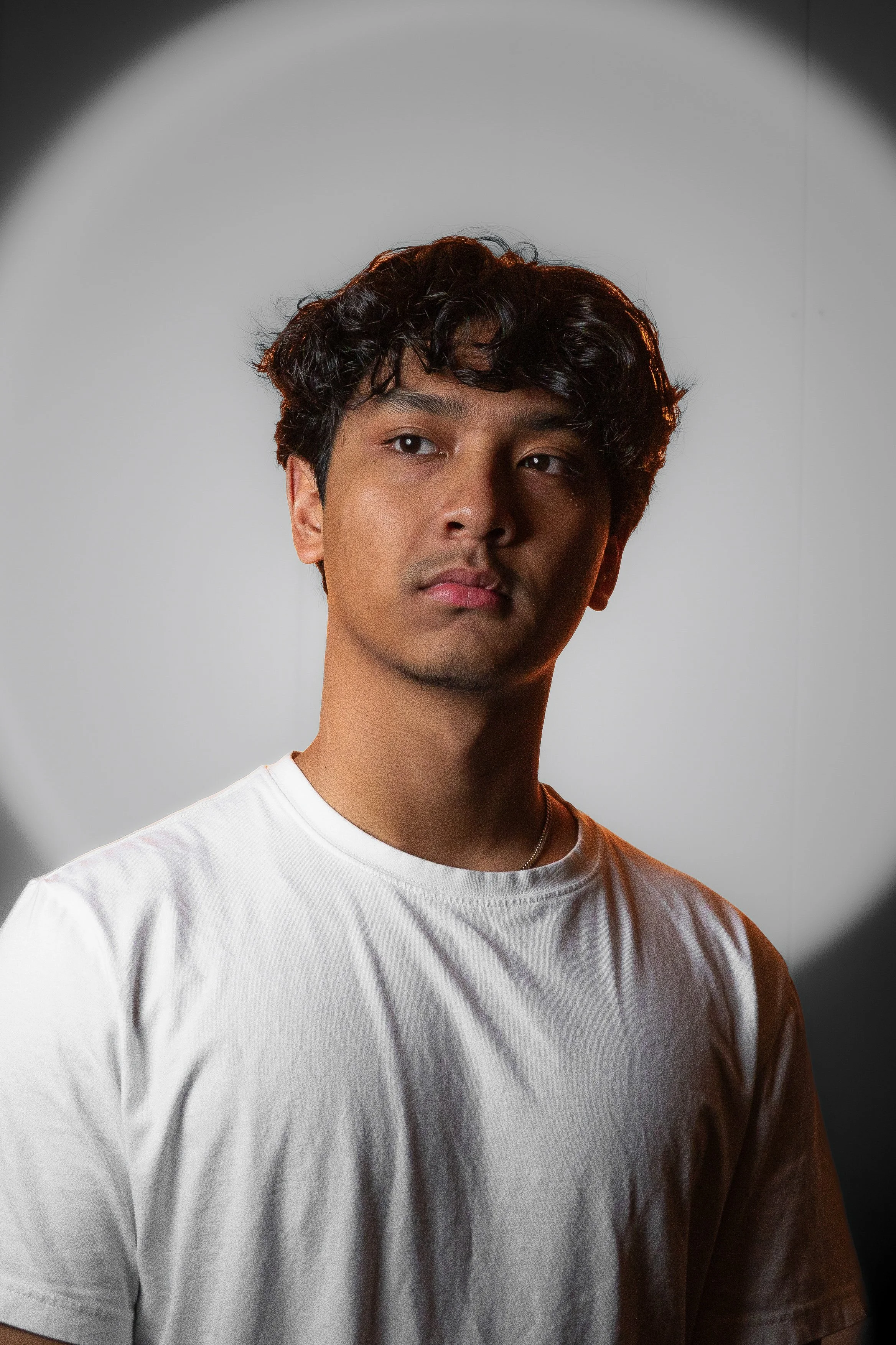 Portrait of a young man with curly dark hair, wearing a white t-shirt, standing against a plain background, lit with dramatic spotlighting.
