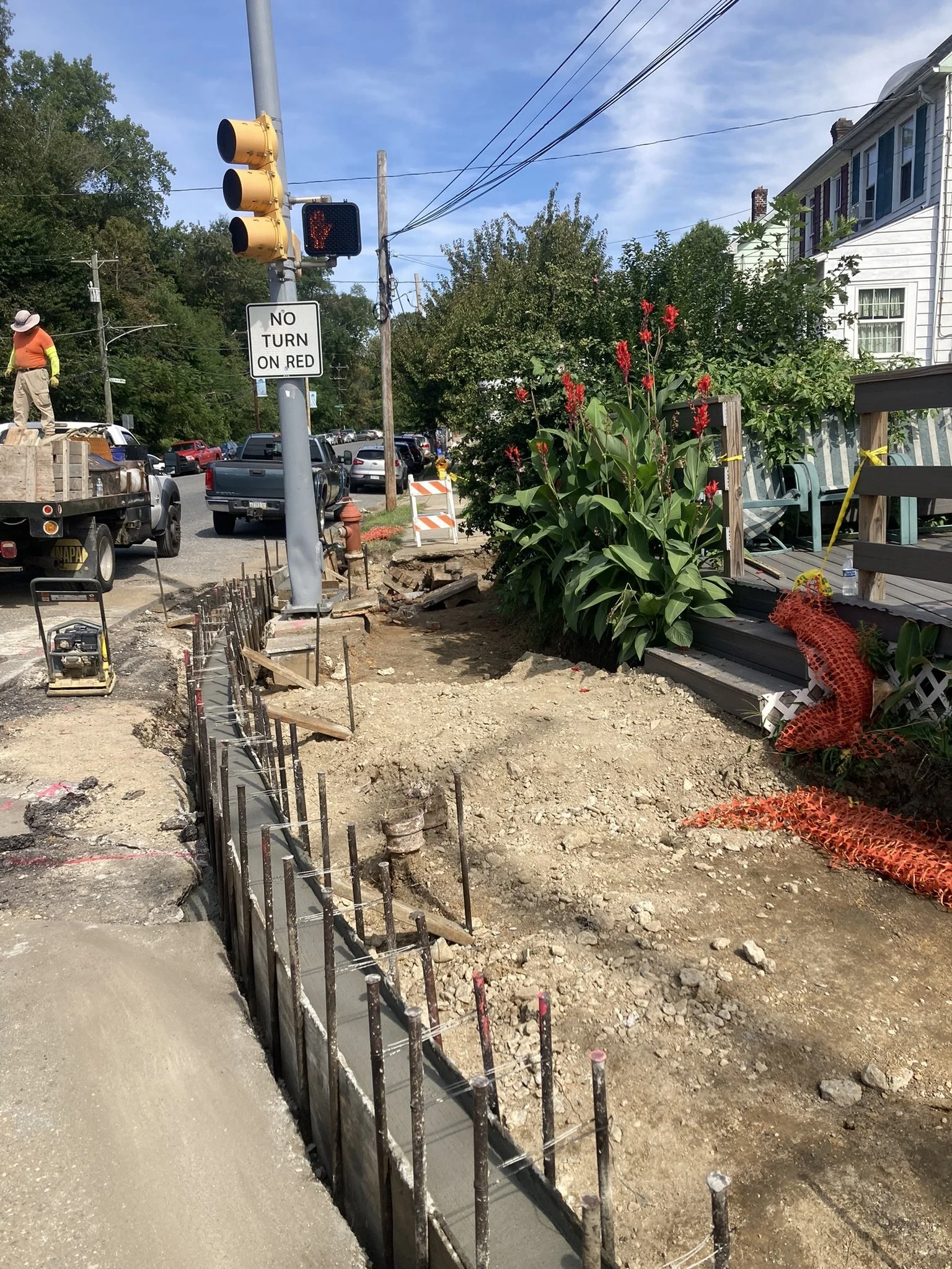 Street construction site with dug-up sidewalk, construction workers, and traffic signal showing red hand. There are red flowers and a white house in the background.