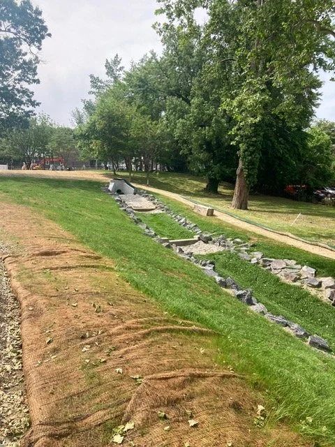 A park with a grassy area and a winding stone path under construction, surrounded by trees.