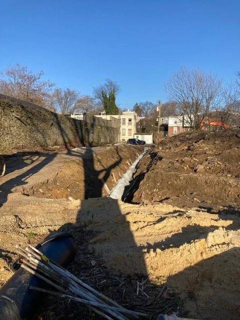 Construction site with a trench and some white piping, with houses and trees in the background under a clear blue sky.