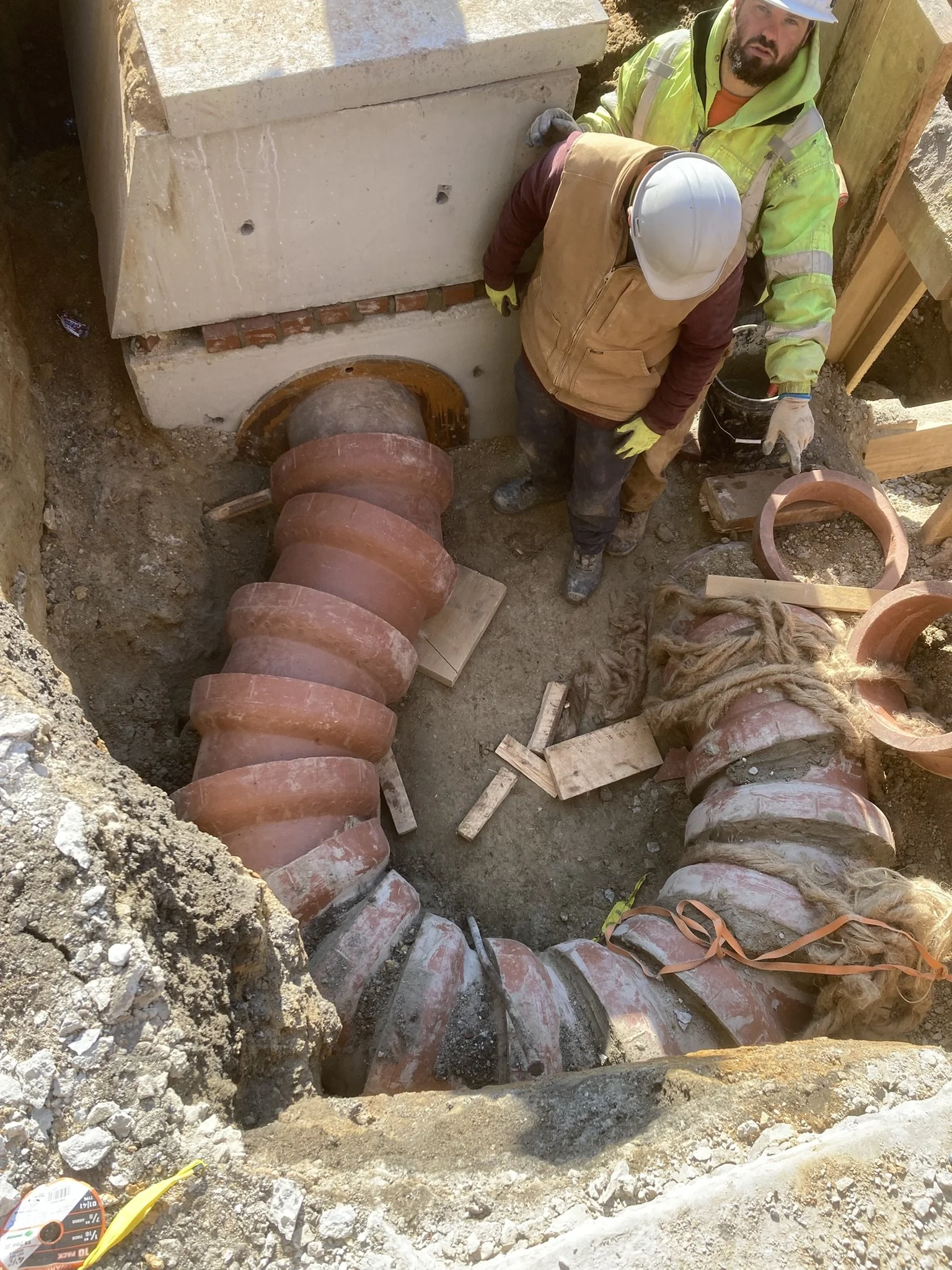 Two construction workers in safety gear installing large curved pipes in an excavation site.
