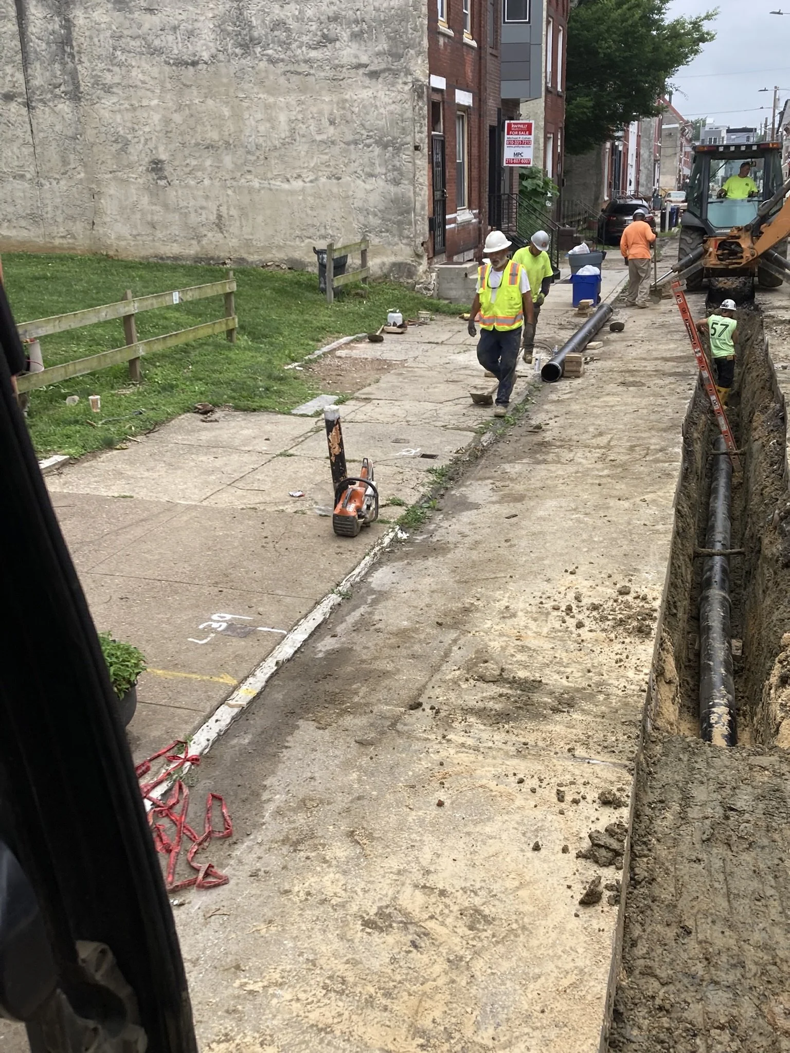 Construction workers in safety vests and helmets working on sidewalk repair with pipes and equipment on a city street.