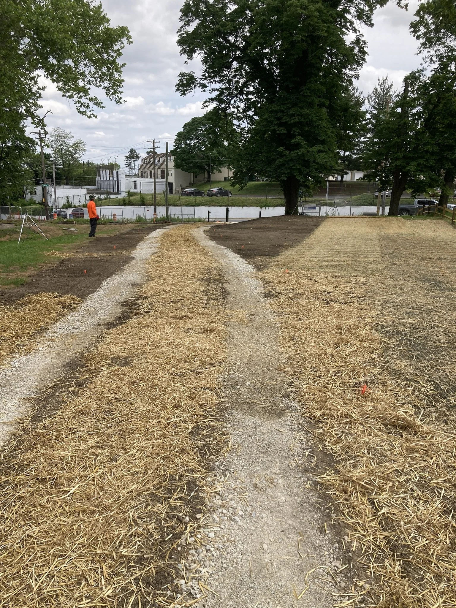 A construction site with a dirt and straw pathway, surrounded by grassy areas and trees, with a worker in an orange vest on the left, and in the background, residential buildings and vehicles are visible.